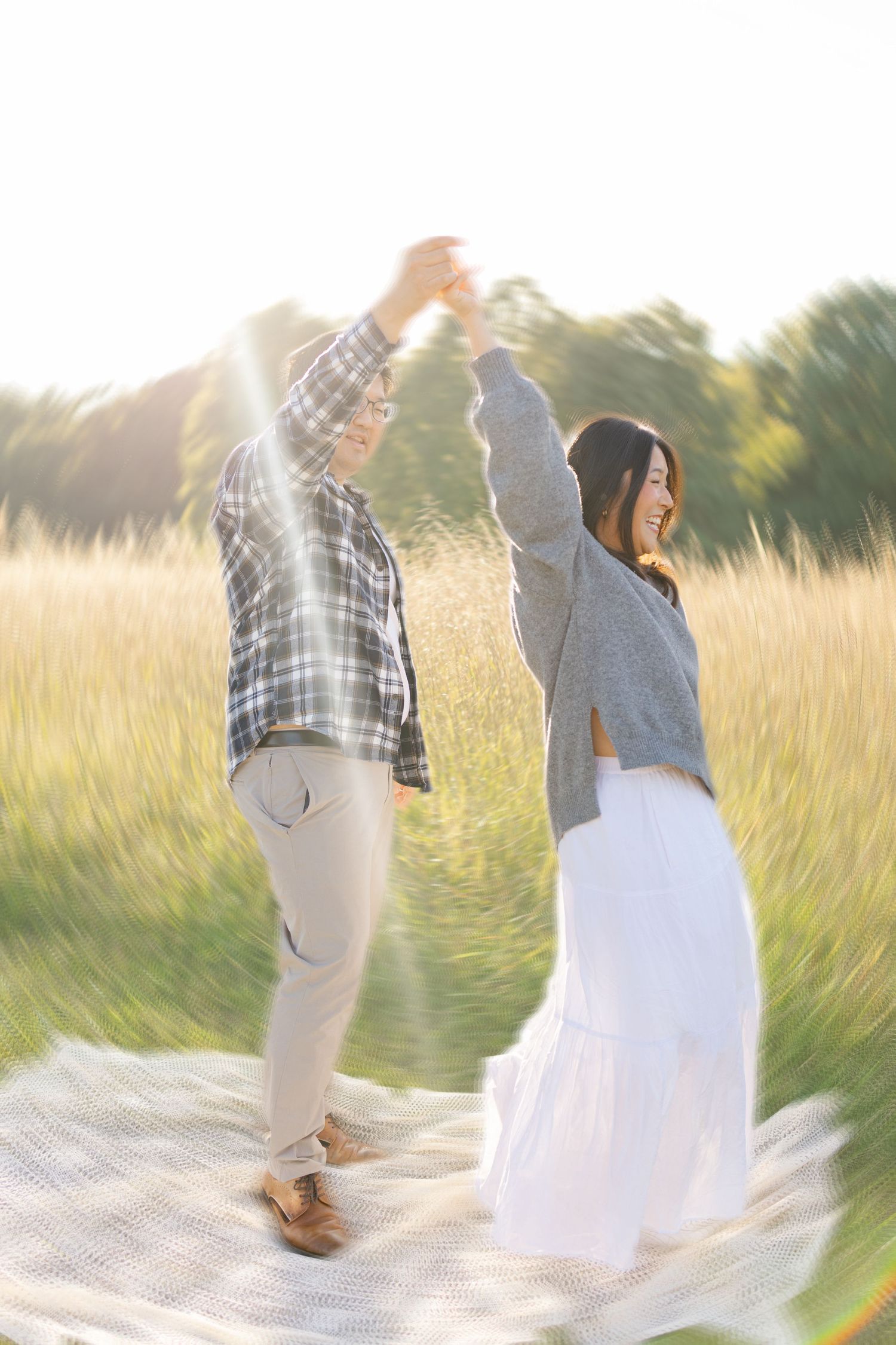 A joyful couple dances together in a sunlit field, the woman in a white skirt laughing as they spin hands raised.