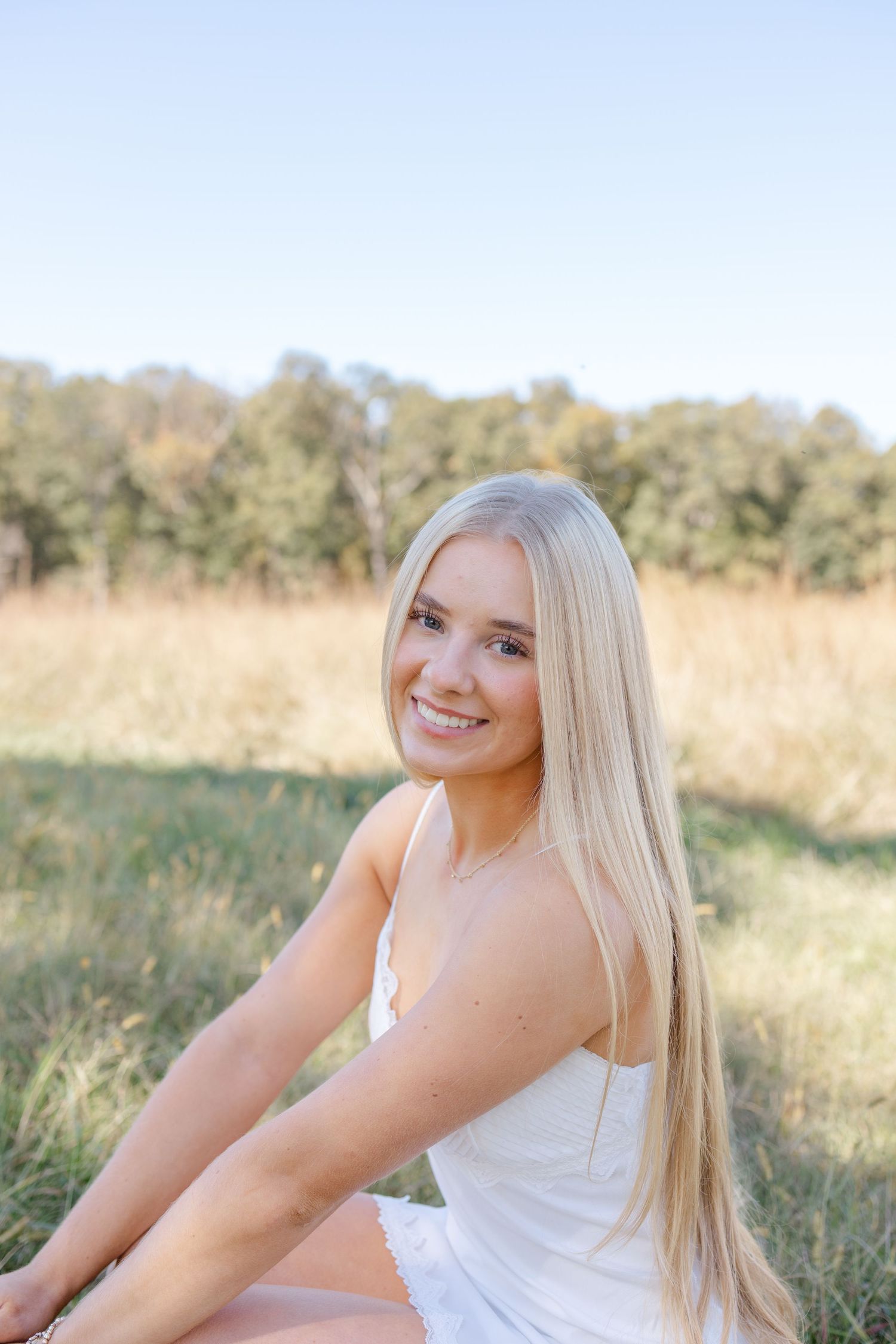 A young woman in a white dress poses in a sunny field with golden grass and trees in the background.