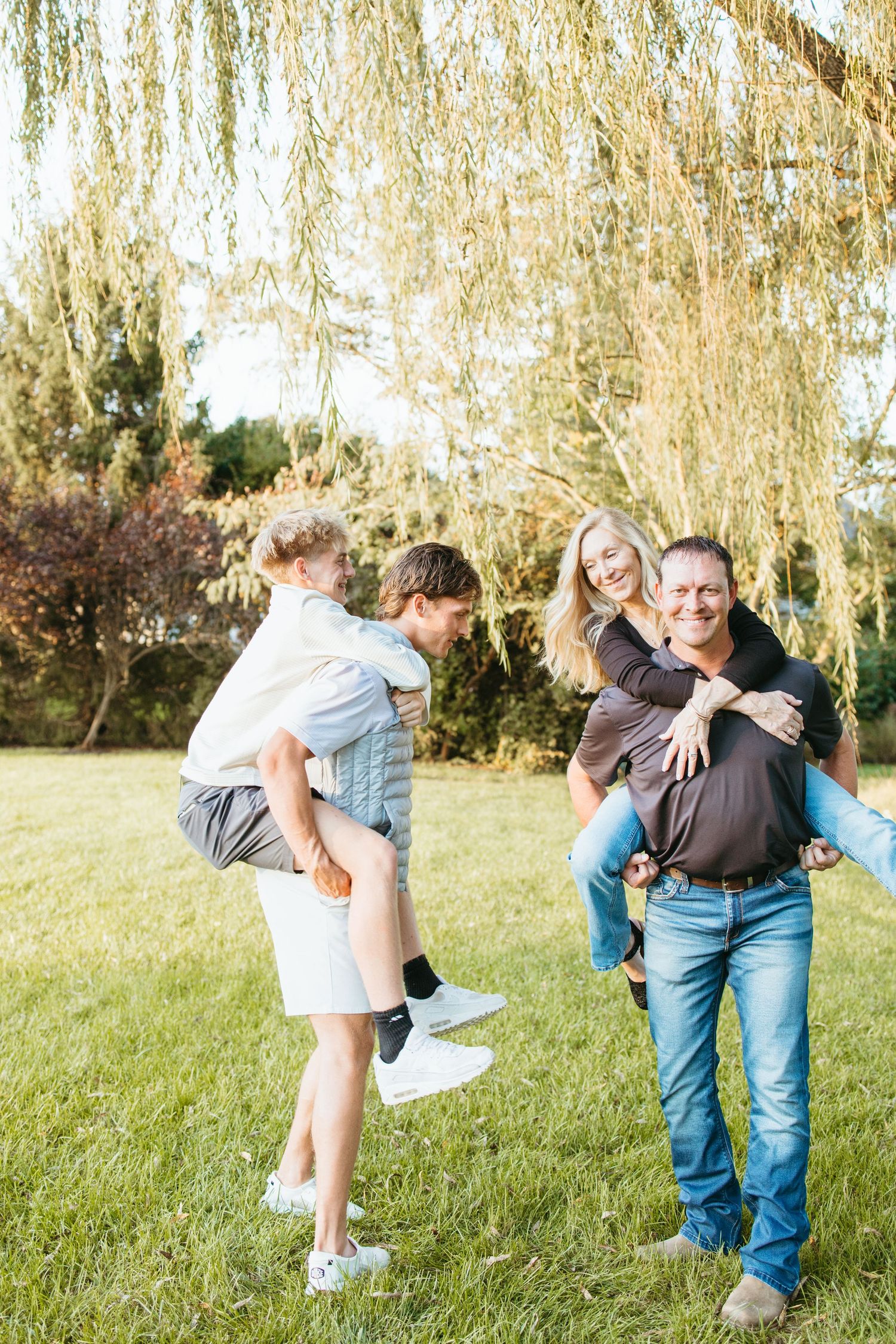 Family enjoying piggyback rides outdoors on green grass under a weeping willow tree on a sunny day.