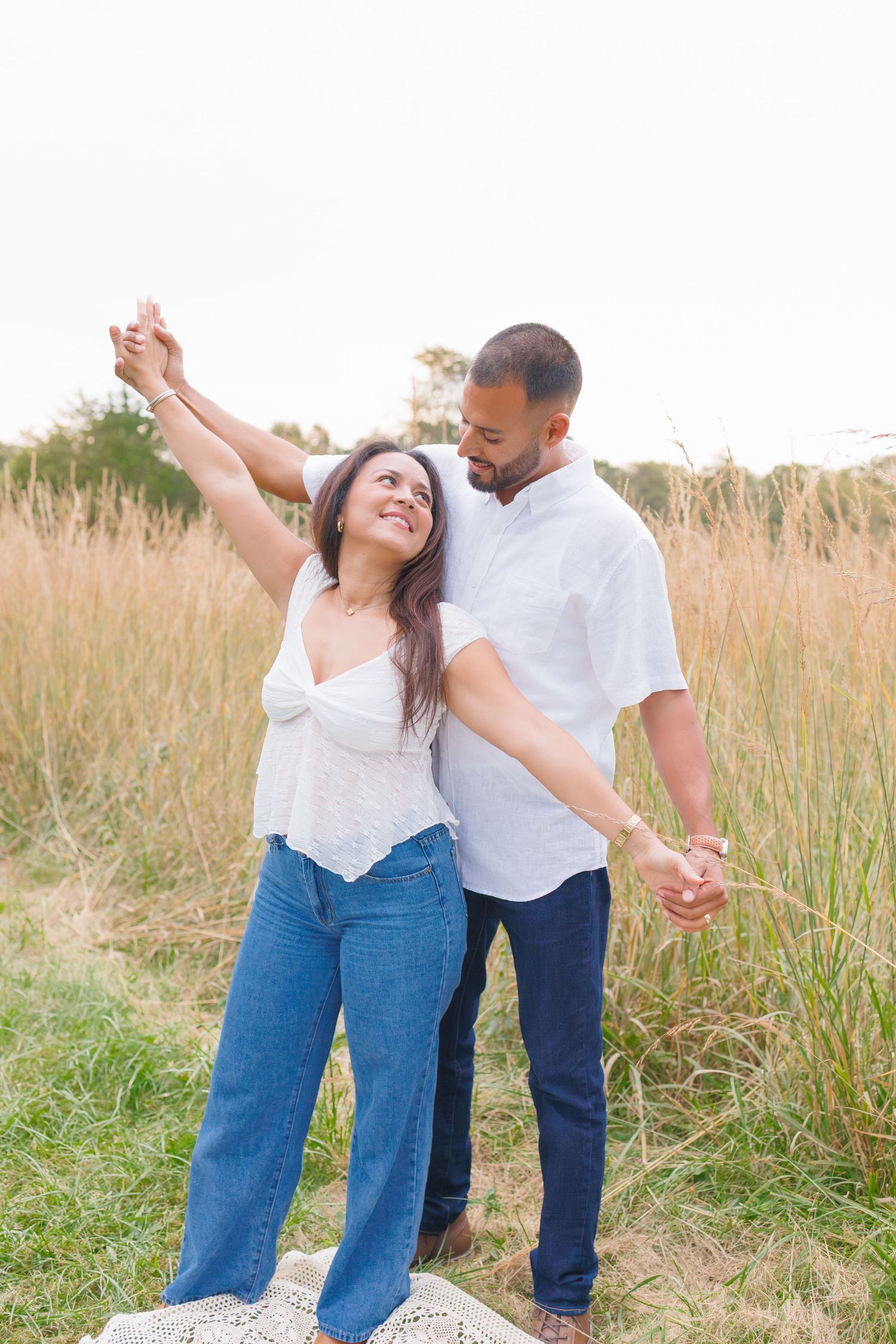Happy couple dancing in a golden field, woman raising arm joyfully, both wearing white tops and blue jeans.