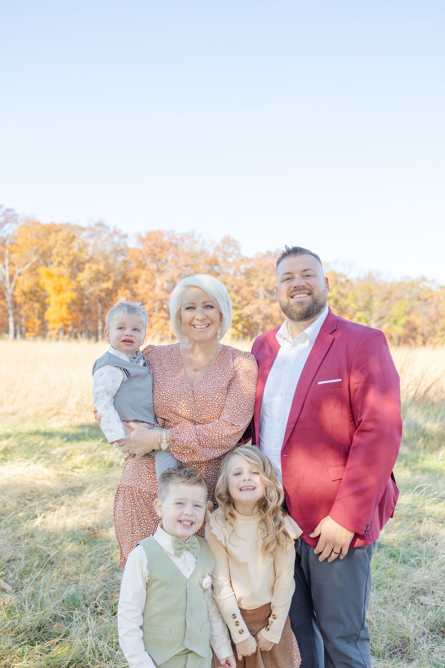 Family of five posing outdoors in fall field, woman holding toddler, man in red blazer, two young children smiling in front.