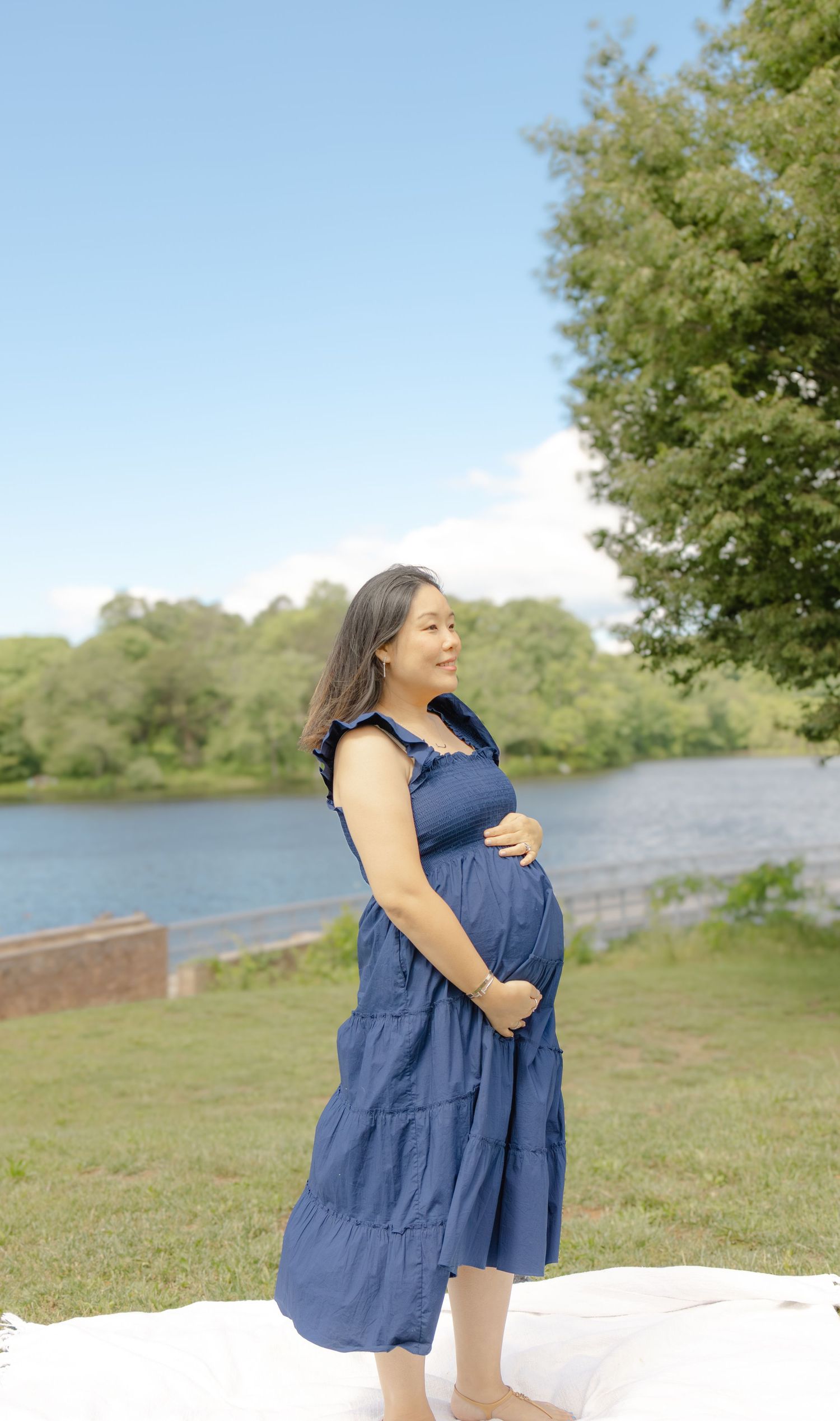 Pregnant woman in blue tiered dress cradling her baby bump outdoors by a serene lake surrounded by lush green trees.