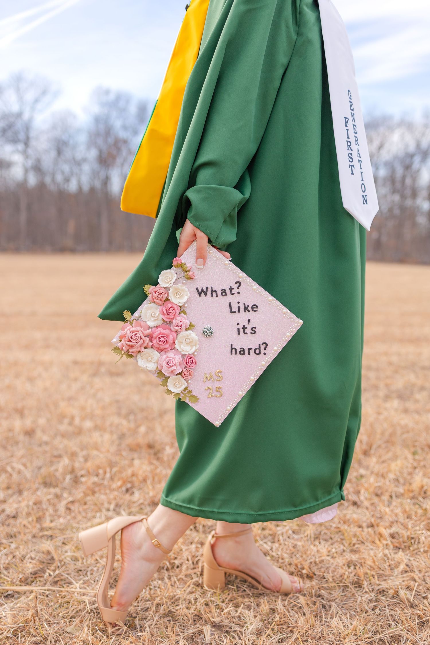 Graduate in green gown holds decorated cap reading What Like its hard with floral accents, standing in field wearing heels.
