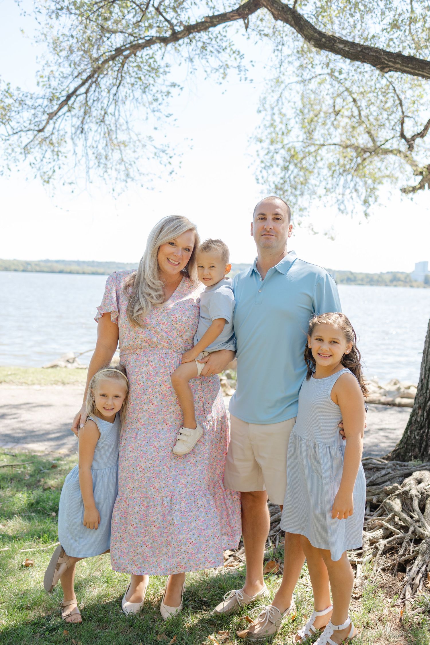 Family in coordinated pastel outfits poses together near a waterfront on a sunny day.