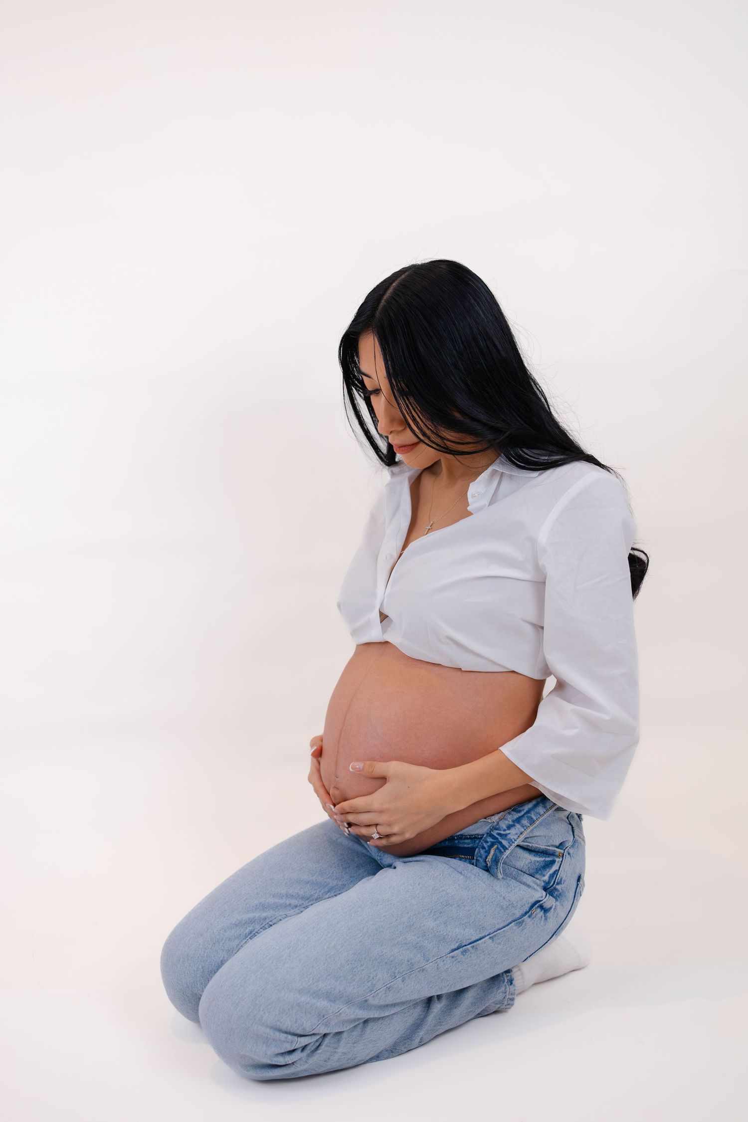 Pregnant woman in white shirt and jeans kneeling, cradling her bare baby bump with both hands.