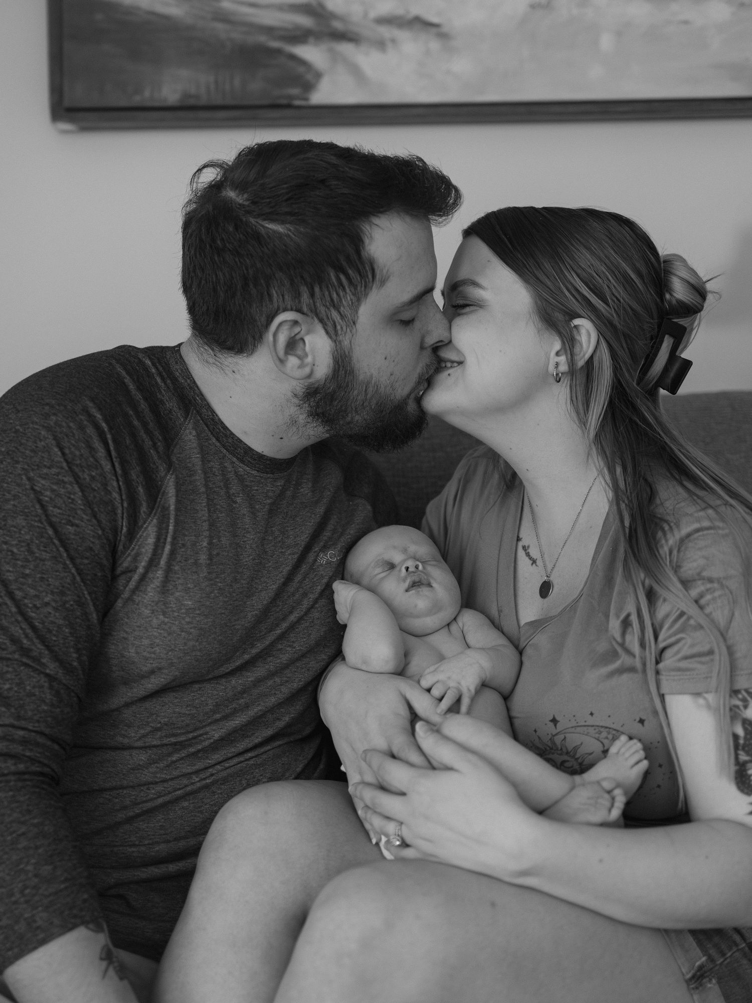 Black and white photo of a couple kissing while holding their newborn baby, capturing a tender family moment.