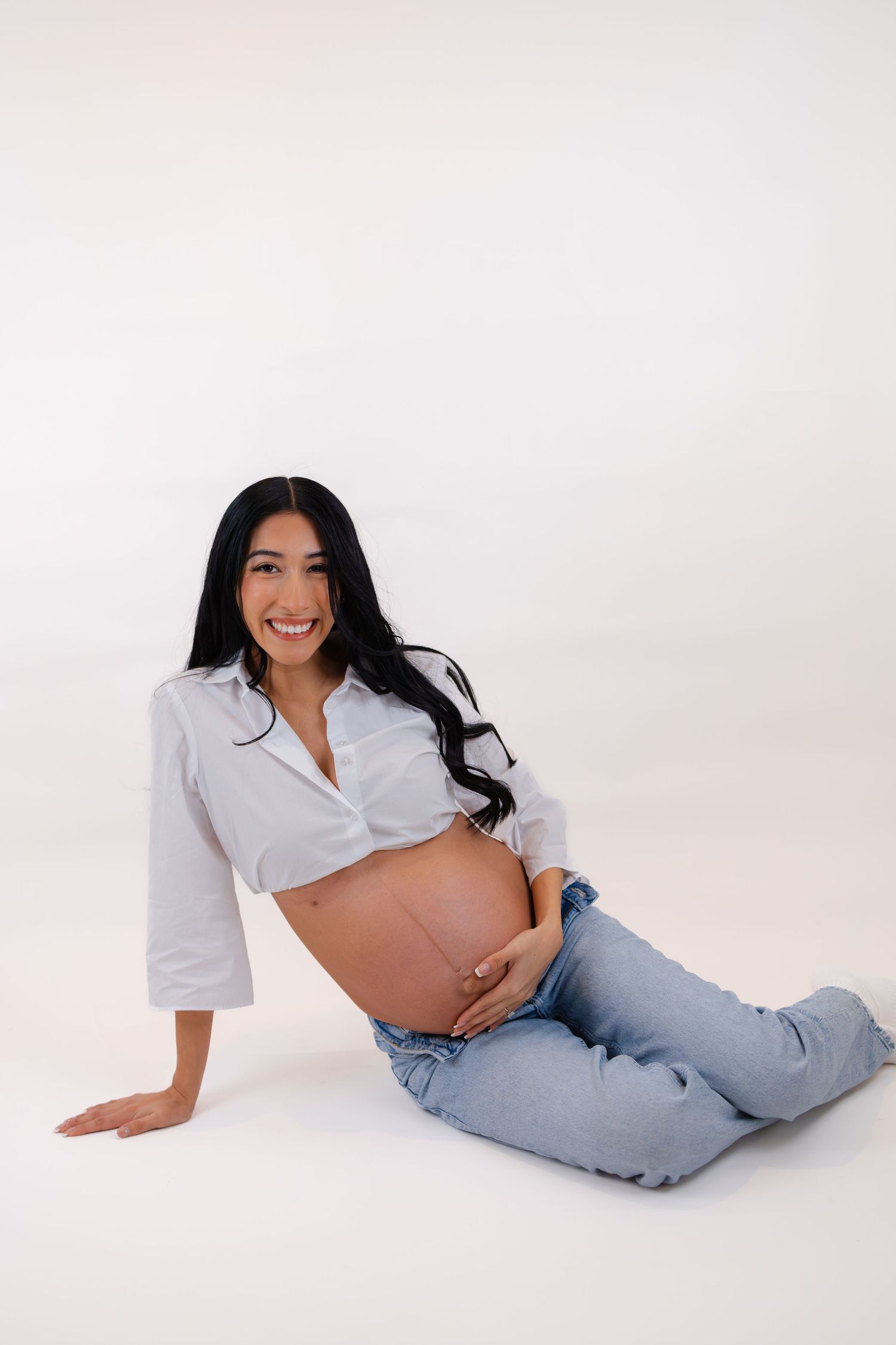 Smiling pregnant woman in white shirt and jeans sitting on floor, cradling her baby bump against white background.