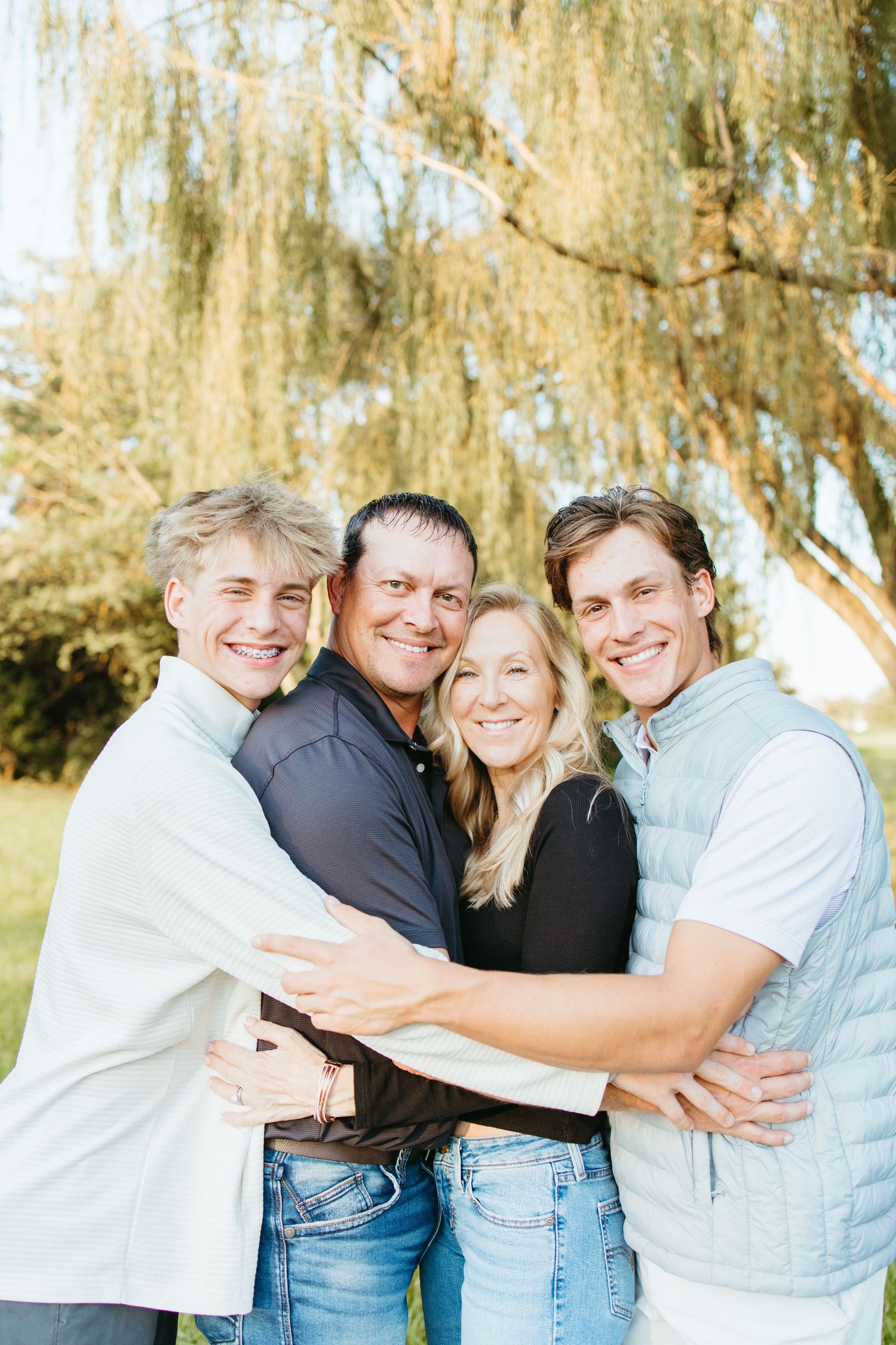 Happy family of four hugging outdoors near a willow tree, smiling together in casual attire during a sunny day.
