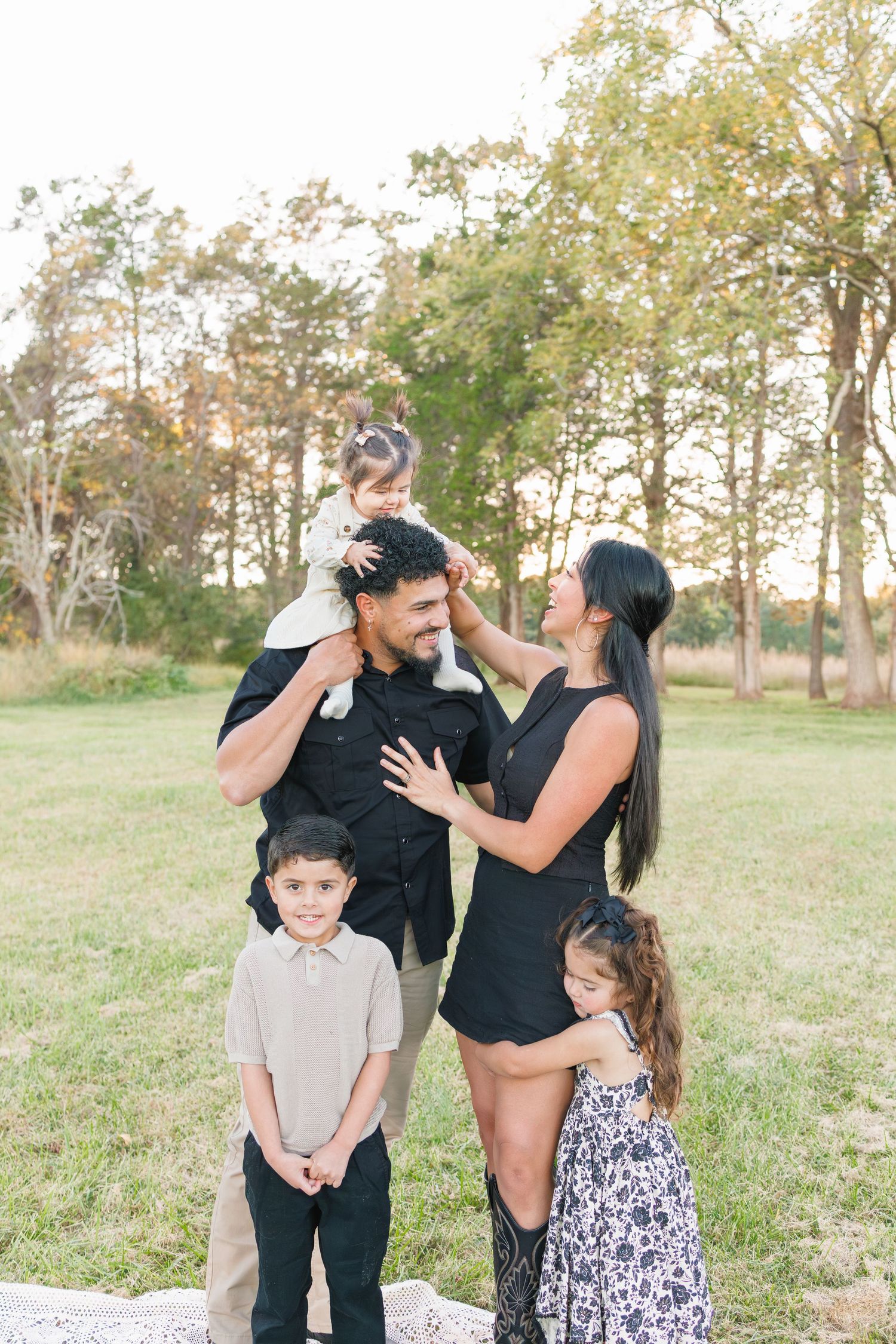 Family of five posing outdoors in a park, parents in black outfits with three young children, golden hour lighting.