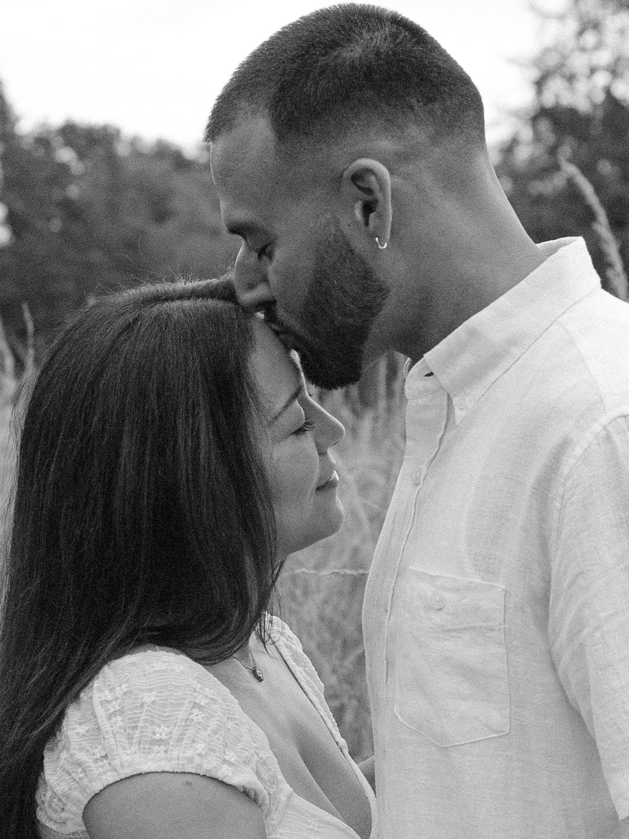 Black and white photo of a romantic couple sharing an intimate moment outdoors in a field, man kissing womans forehead.