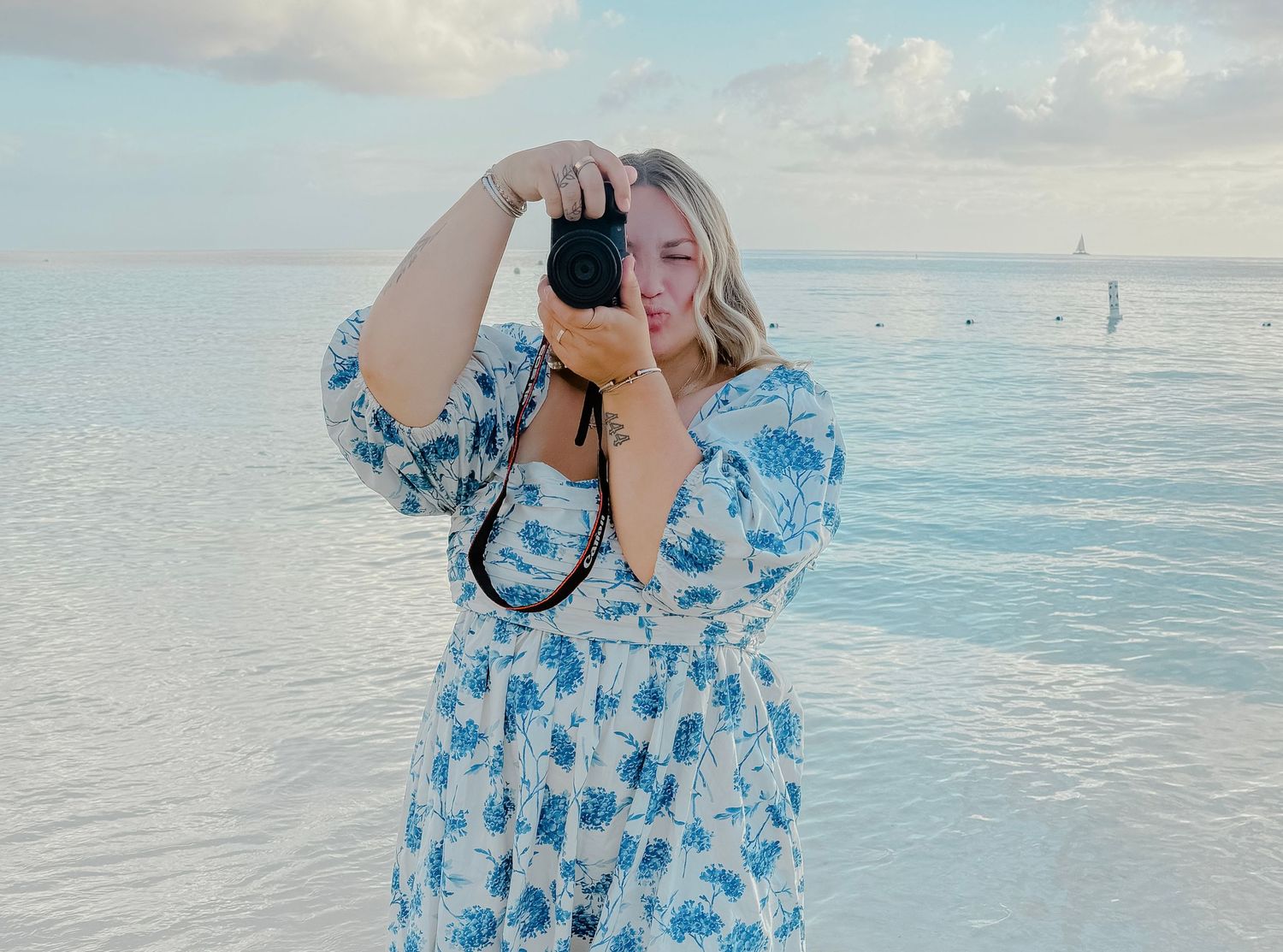 Woman in floral blue dress photographing ocean scenery on a sunny beach with calm turquoise waters.