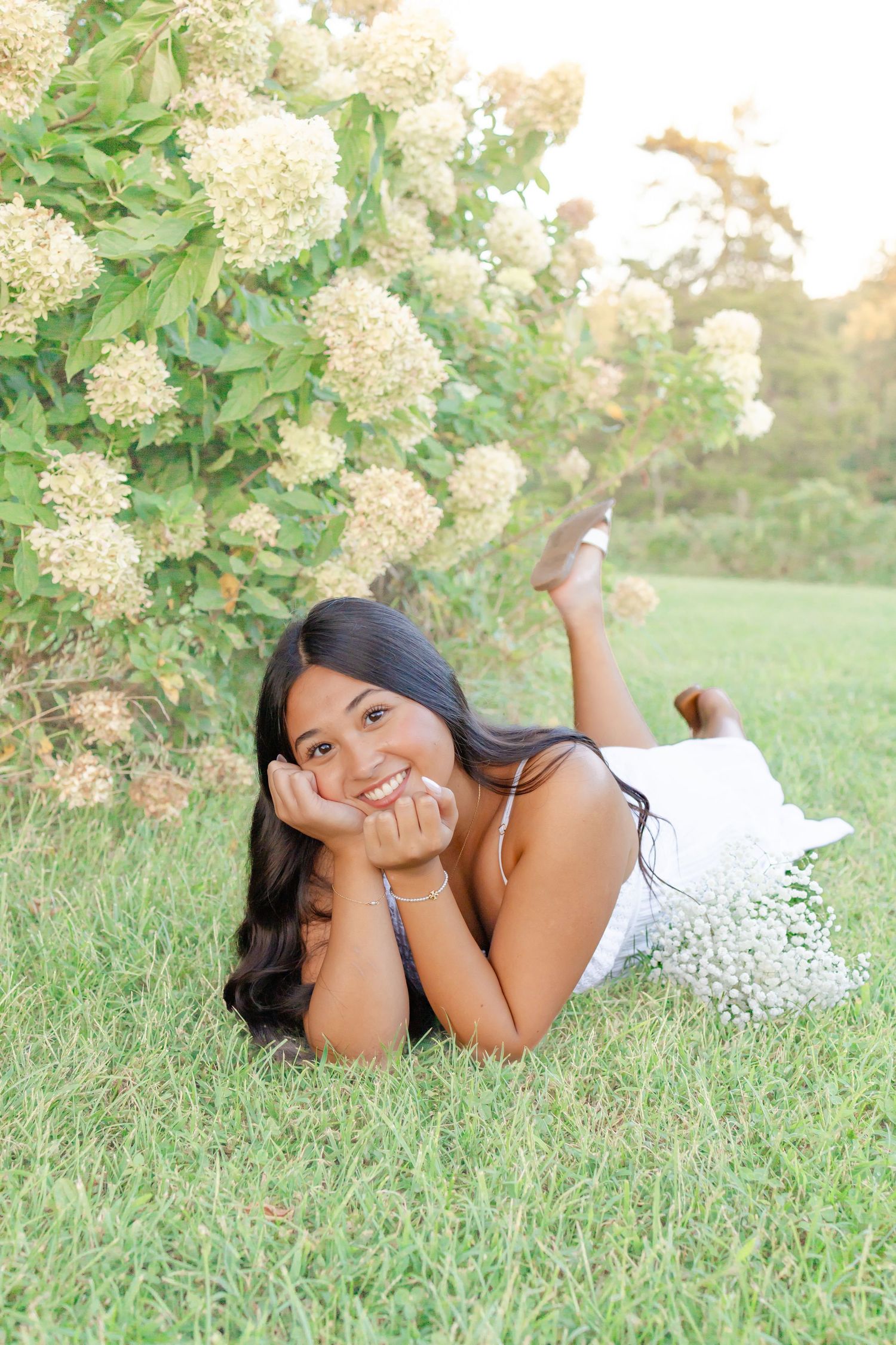 Young woman in white dress lying on grass, smiling, near blooming white hydrangea bushes in a sunny park.