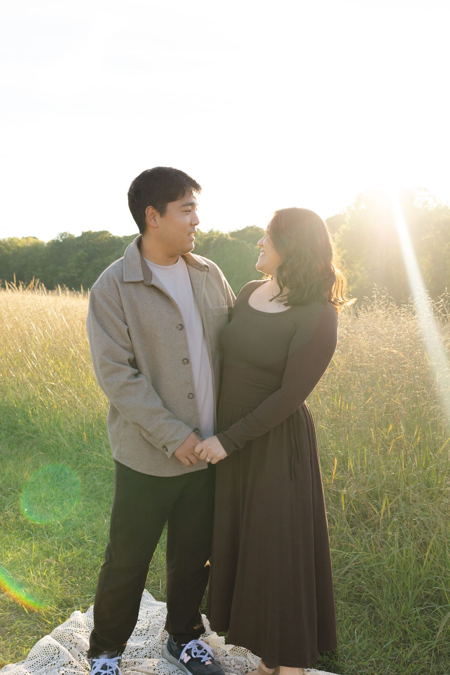 Couple holding hands in a sunlit field at golden hour, man in beige jacket and woman in olive dress.