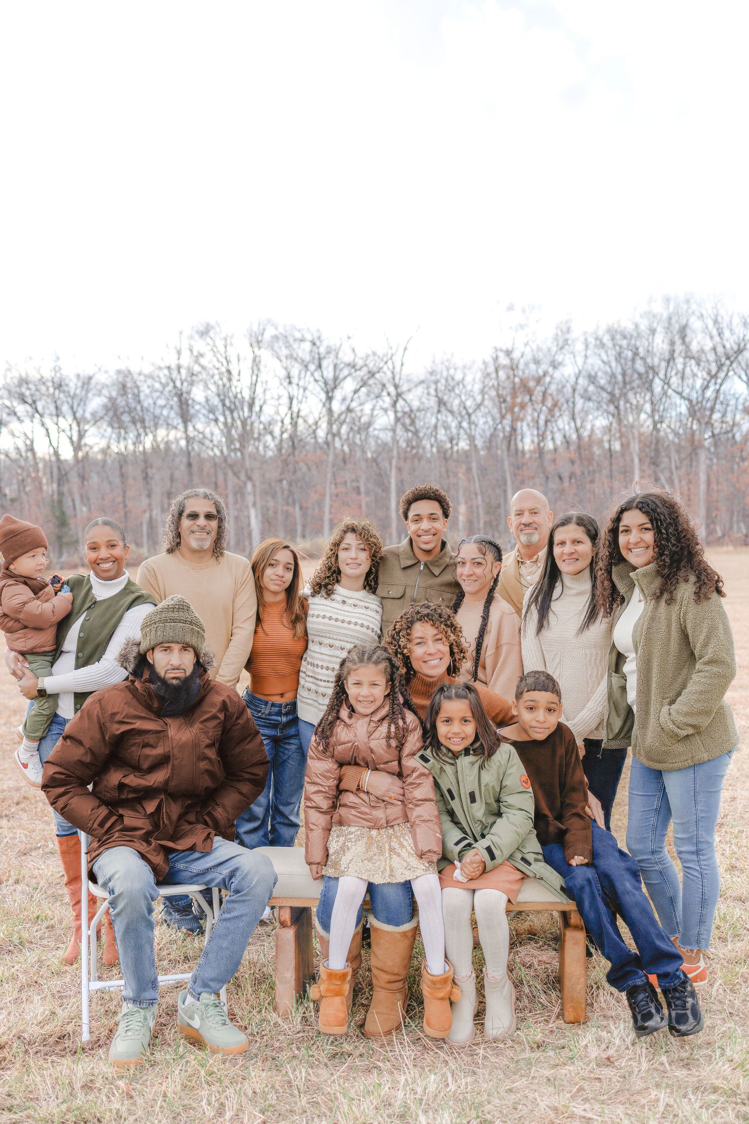 Large multi-generational family posing outdoors in autumn, wearing earth-tone winter clothing, with bare trees in background.