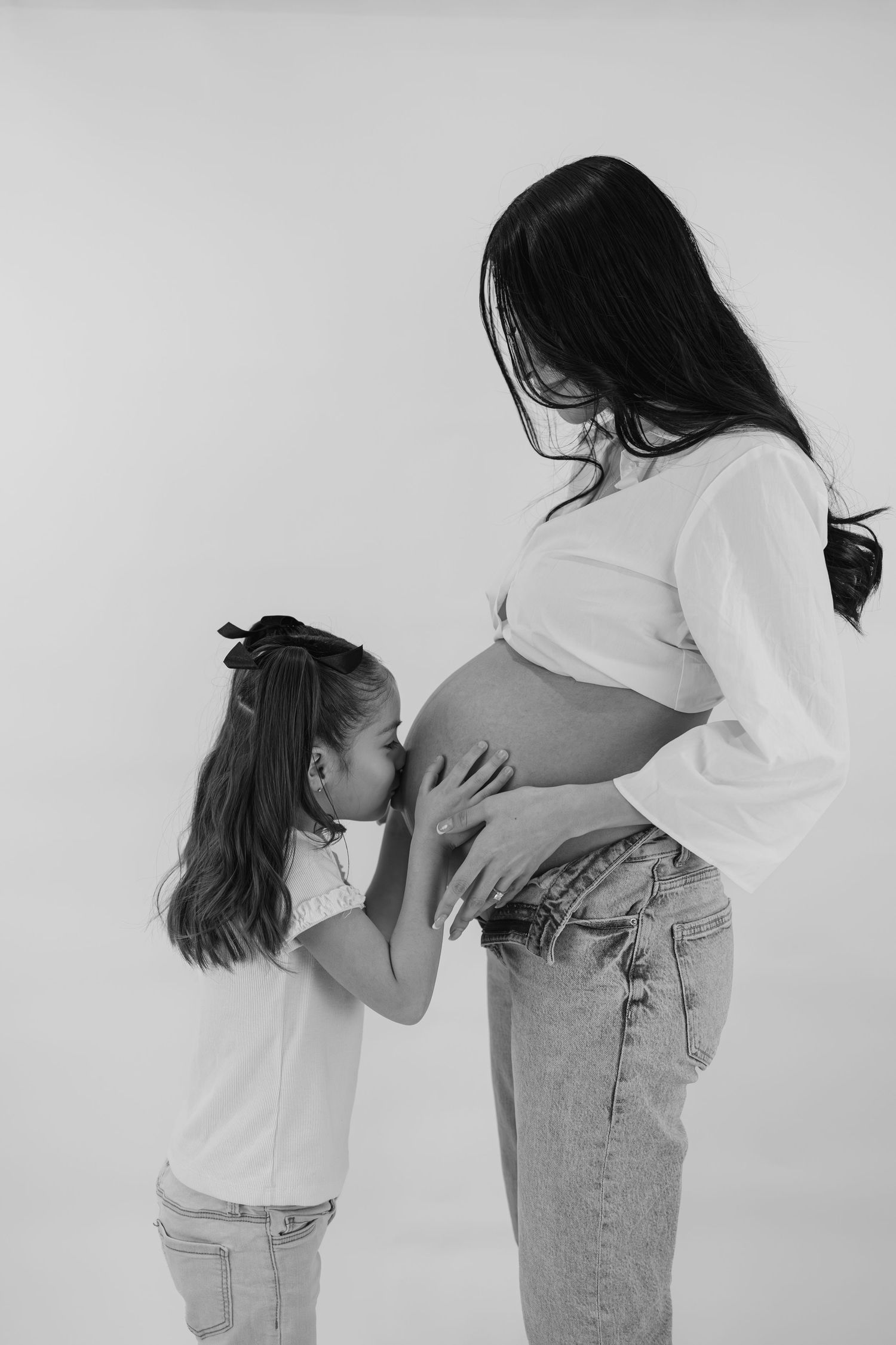 Black and white photo of a young girl kissing her pregnant mother's baby bump in a tender maternity portrait.
