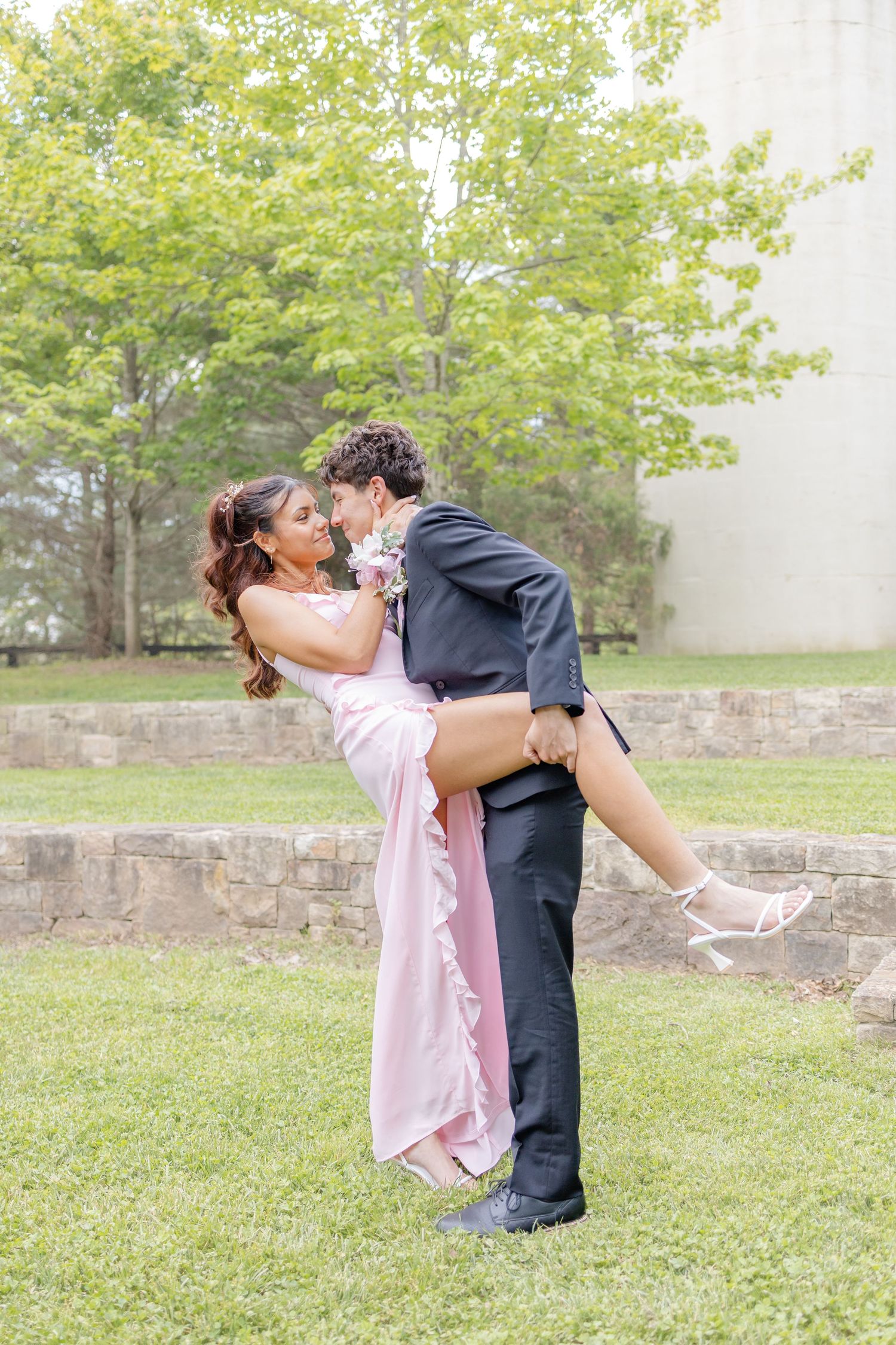 A young couple in prom attire shares a romantic kiss outdoors, the man lifting the woman in a pink gown.