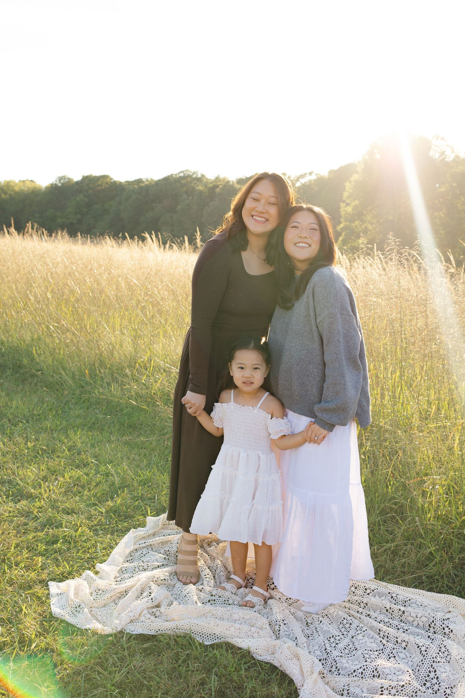 Two women and a toddler girl in white dress pose in sunlit golden field, smiling warmly for a family portrait.