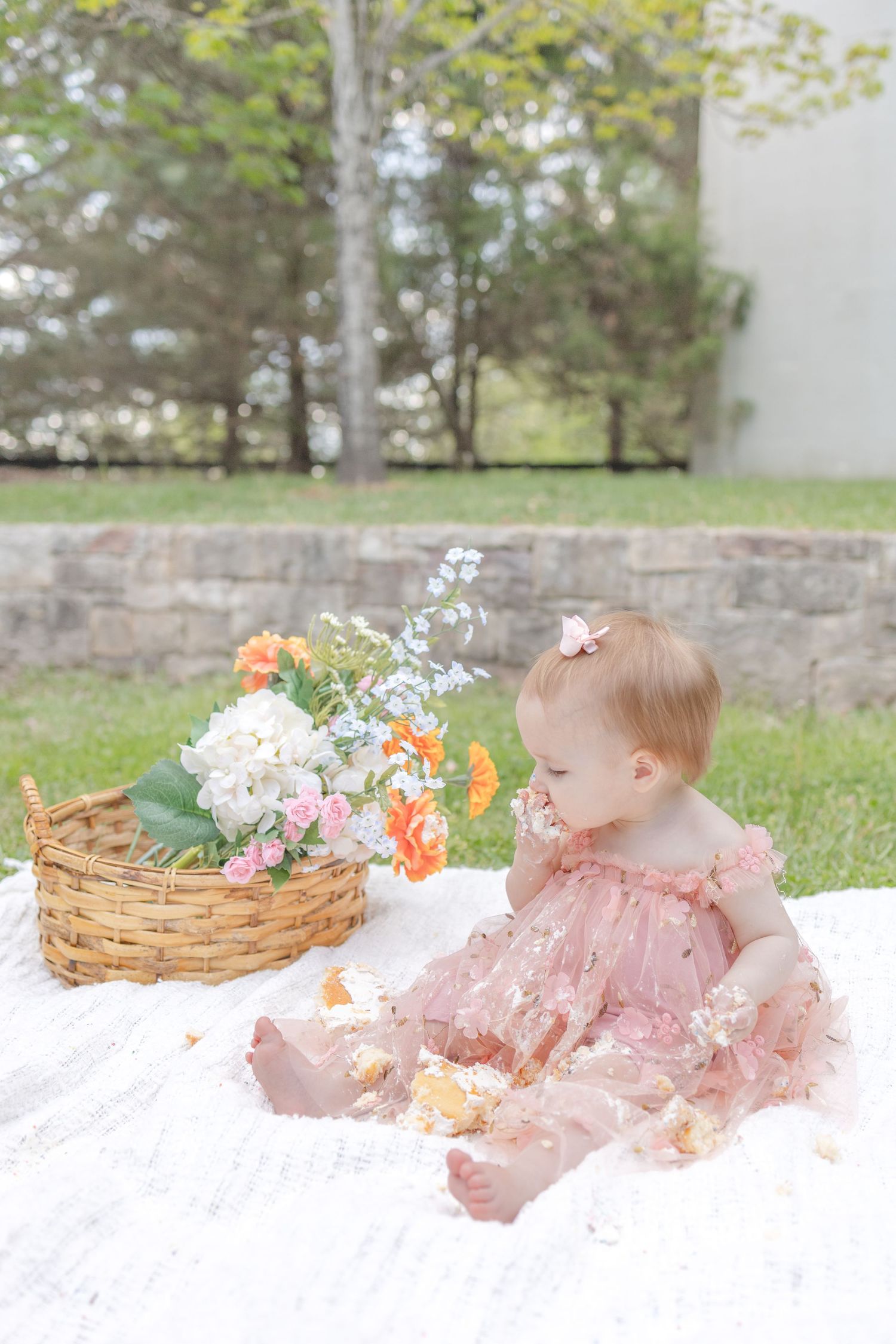 Baby girl in pink floral dress sits on white blanket outdoors near flower basket during first birthday cake smash.