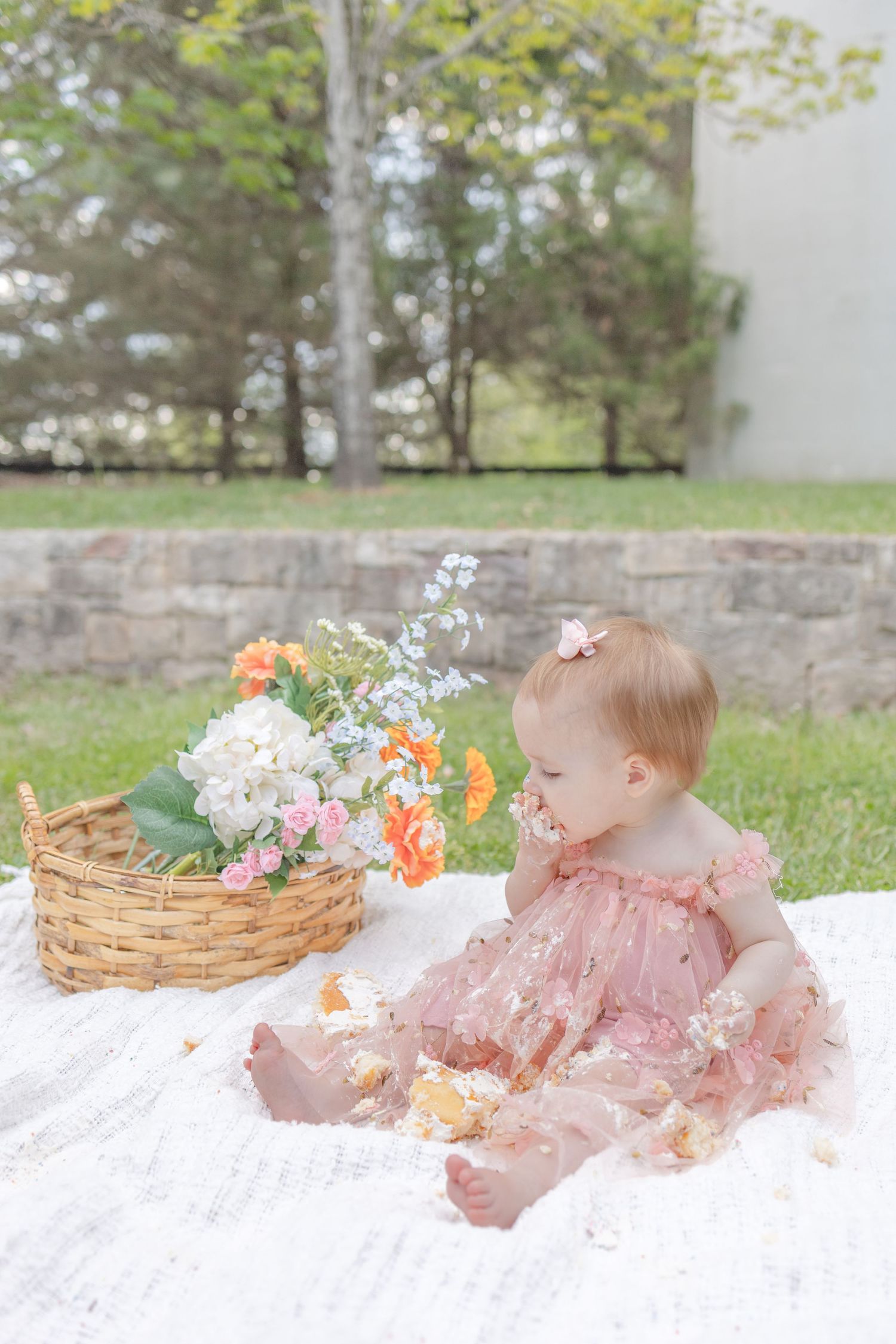 Baby girl in pink dress sits on white blanket beside flower basket outdoors during first birthday cake smash.