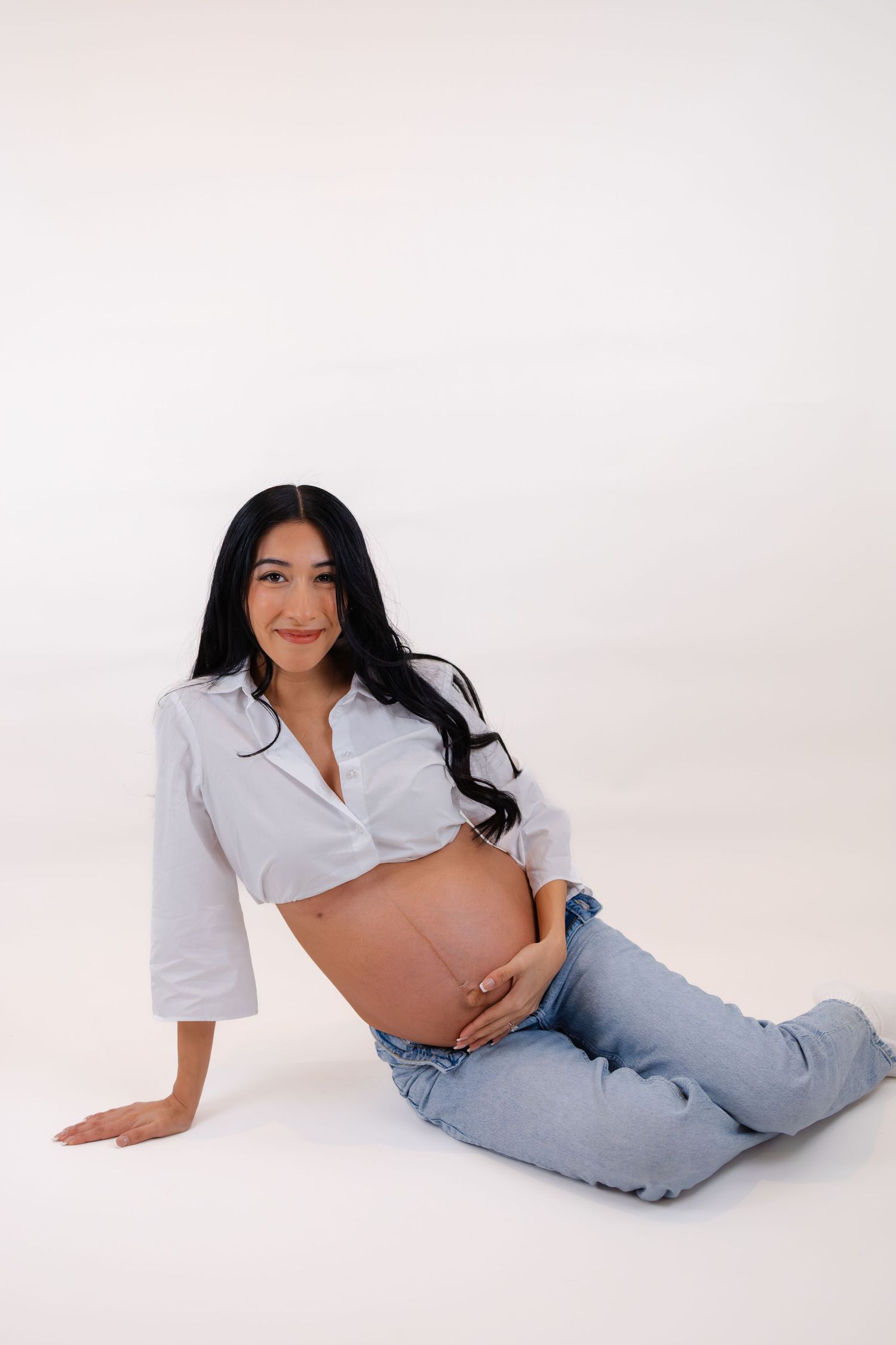 Pregnant woman sitting on floor in white shirt and jeans, cradling bare baby bump, smiling at camera against white background.