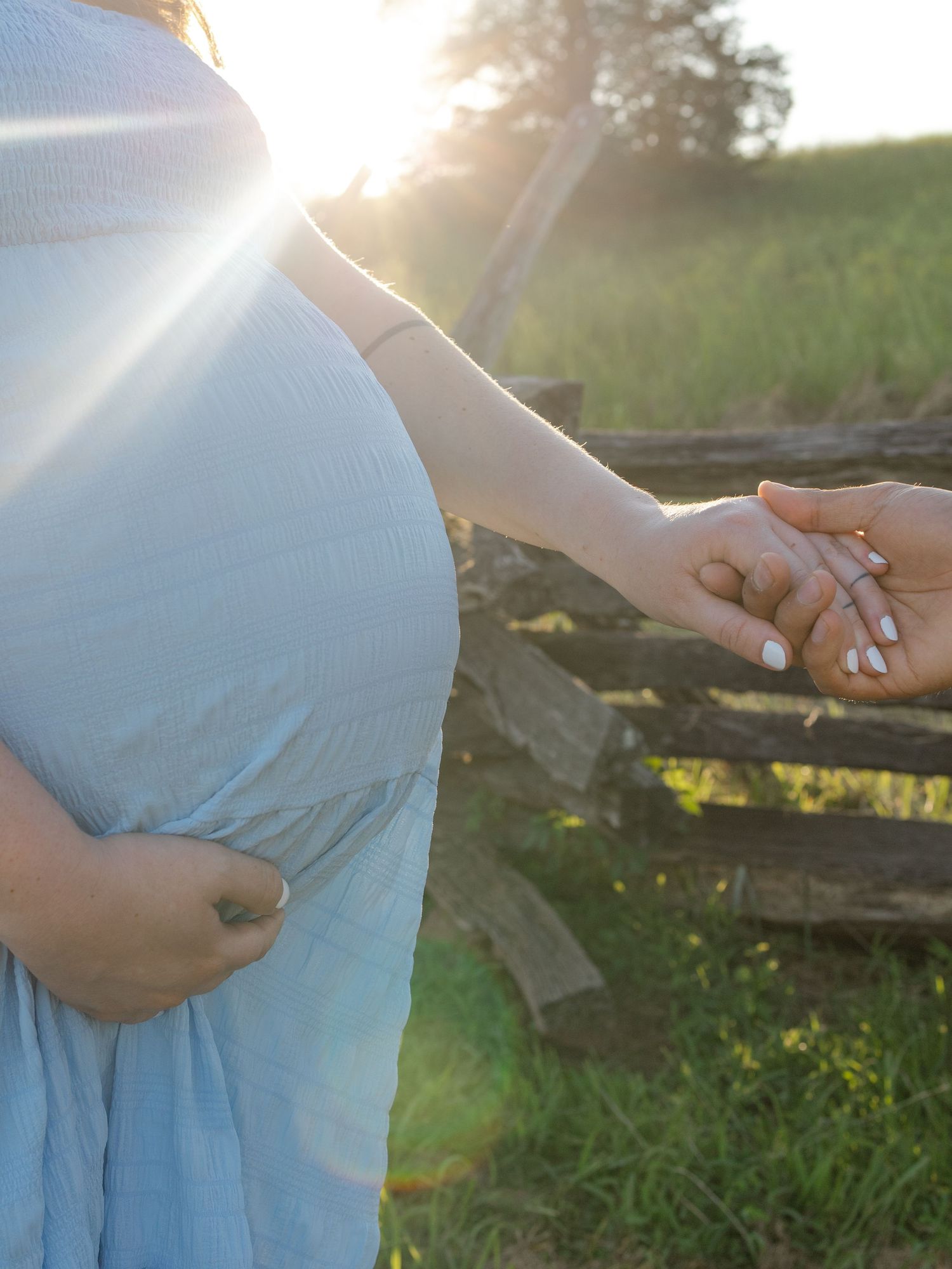 Pregnant woman in blue dress holding hands with partner outdoors at sunset near rustic wooden fence.