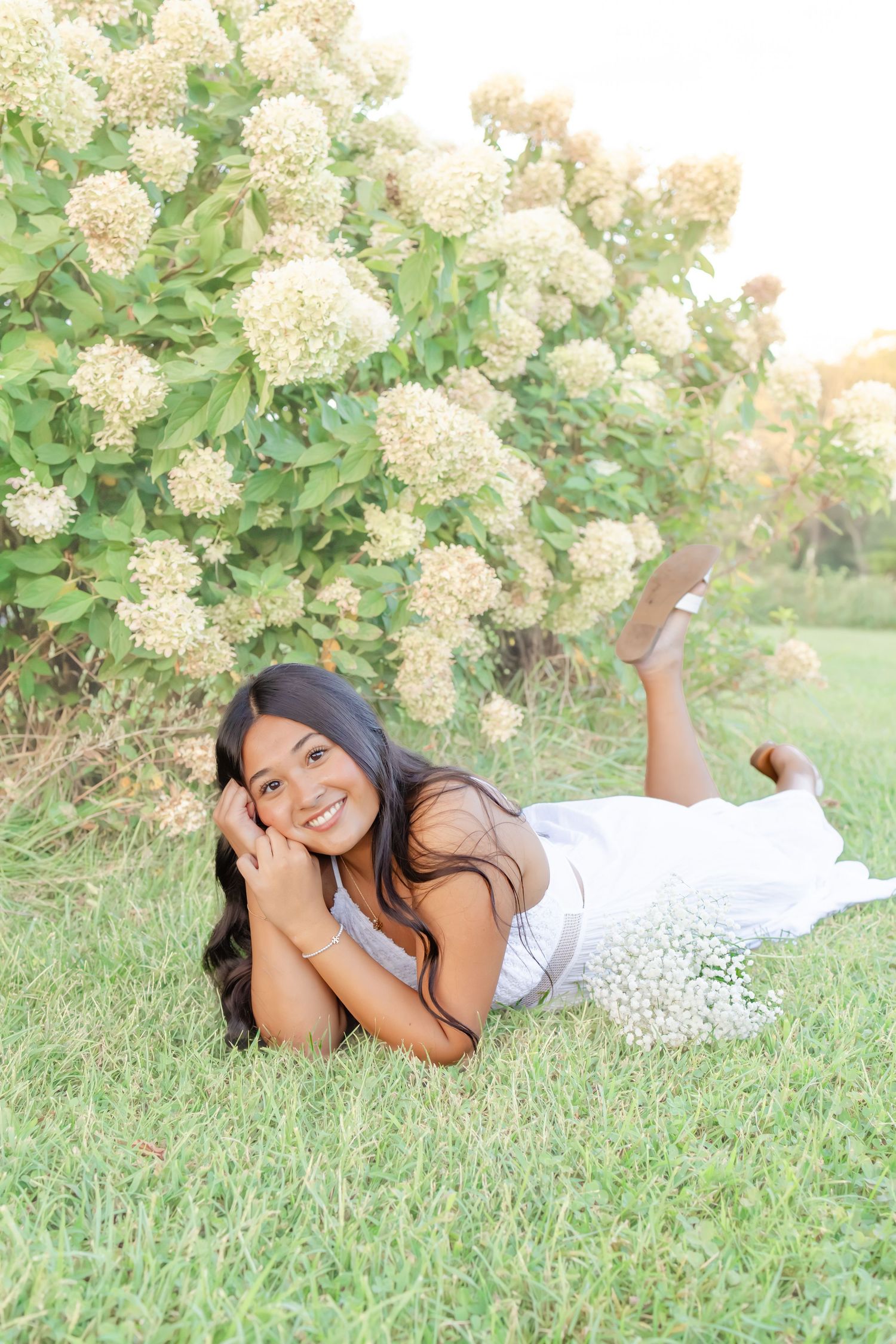 Young woman in white dress lying on grass smiling, surrounded by blooming hydrangea bushes outdoors.