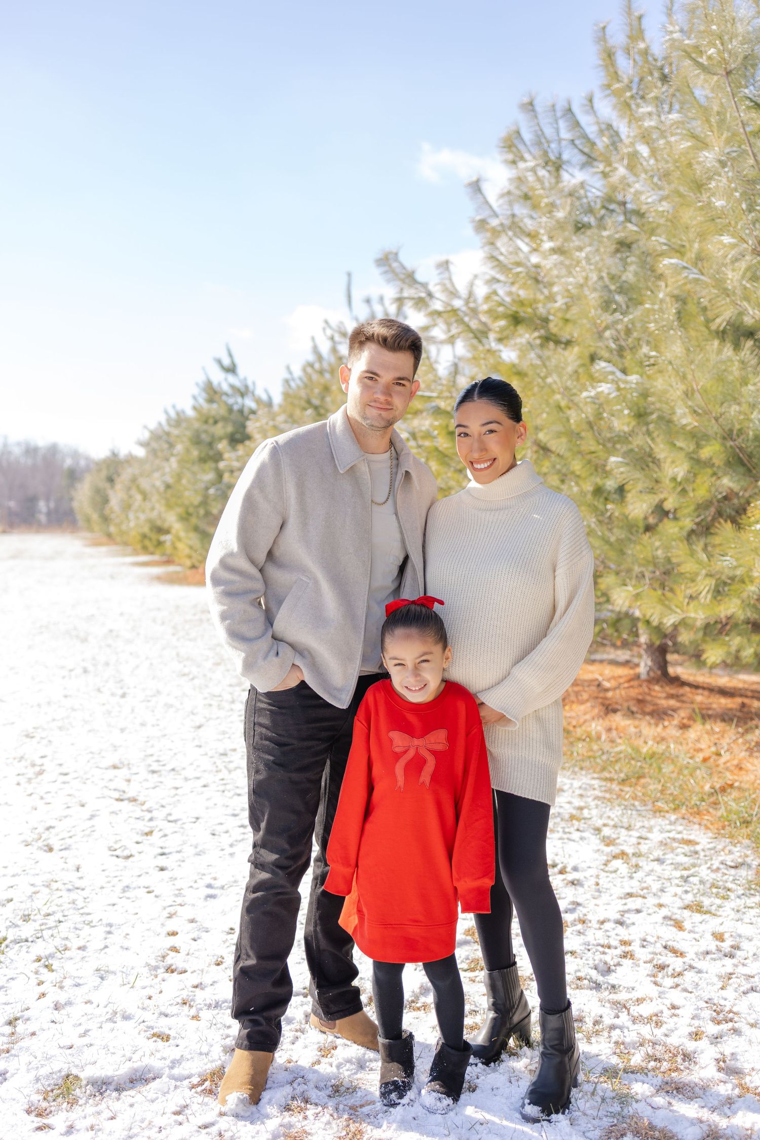 Family of three posing outdoors in winter, parents in neutral sweaters with young girl in red dress, snow-covered ground and pine trees.
