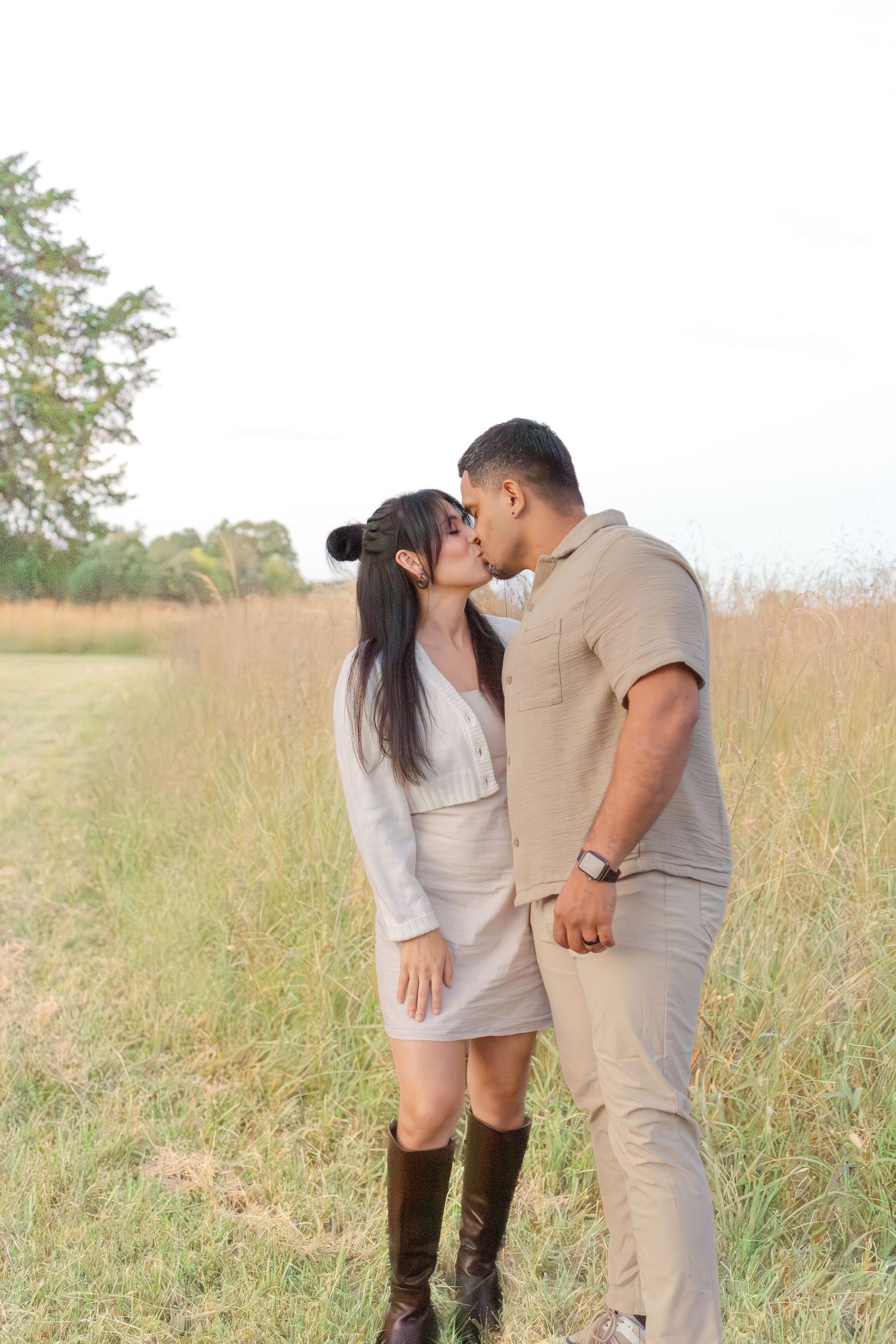 Couple sharing a romantic kiss in a golden grass field, woman in white dress with brown boots, man in beige outfit.