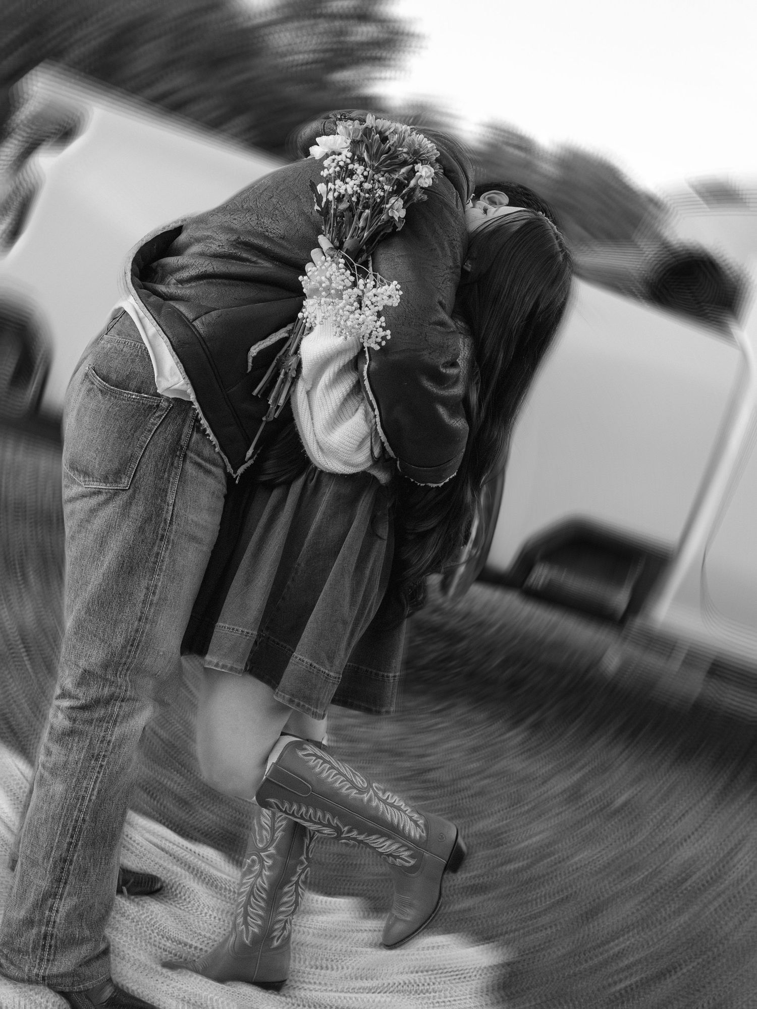 Black and white photo of a couple embracing and kissing, holding wildflowers, wearing jeans and cowboy boots outdoors.