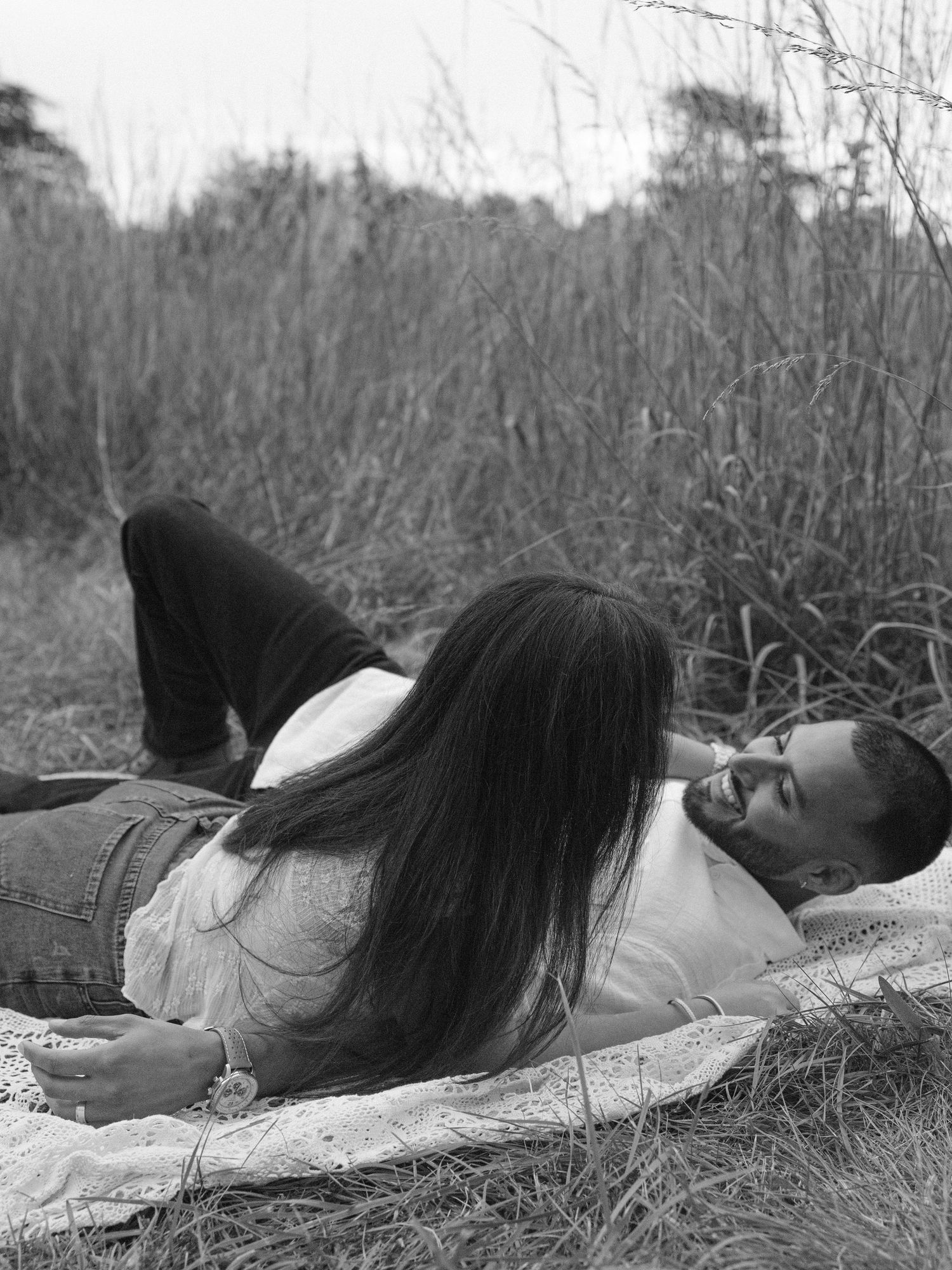 Black and white photo of a couple lying in a grassy field, woman with long hair resting on mans chest smiling.