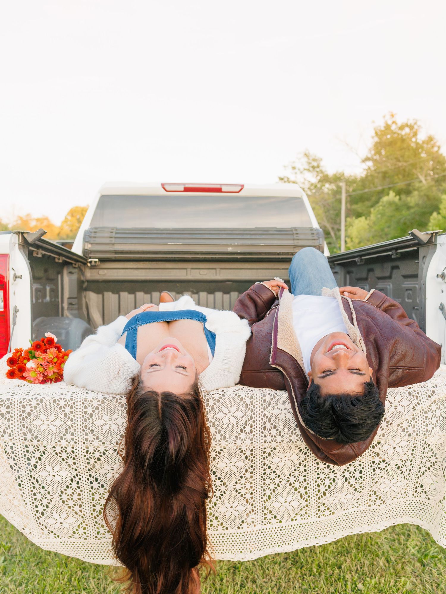 Two people lying on a lace blanket in a truck bed, smiling upward with flowers nearby on a sunny day.