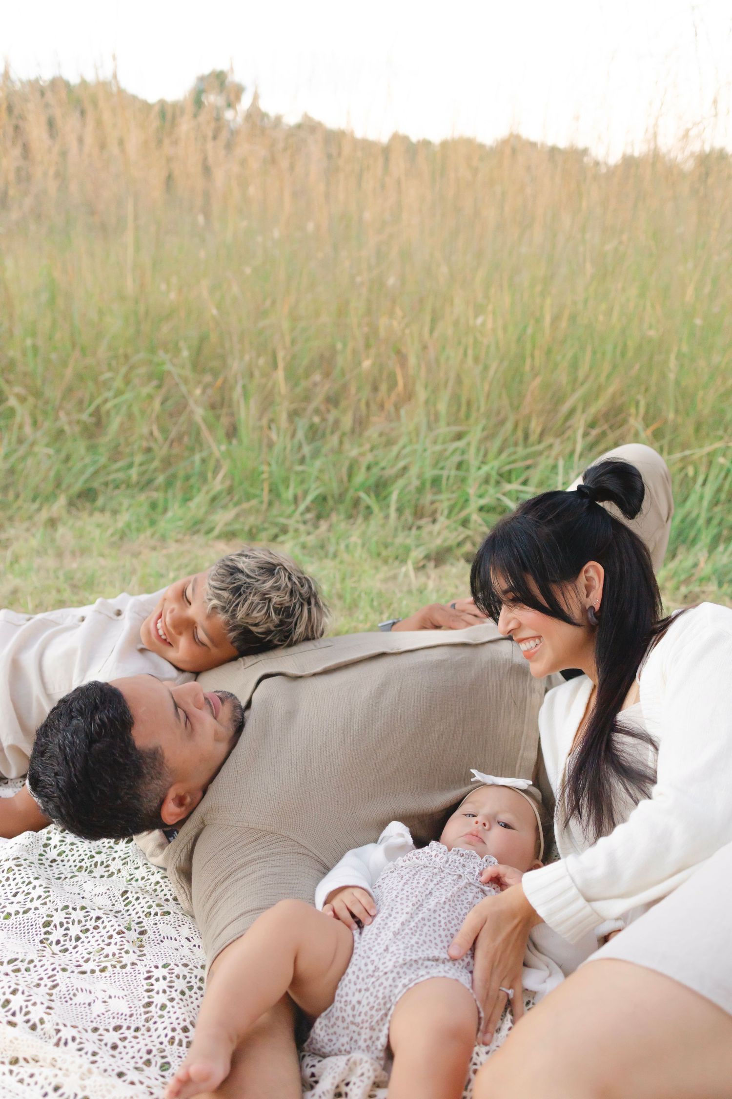 Family relaxing outdoors on blanket in field, parents smiling at children, newborn baby lying between them.