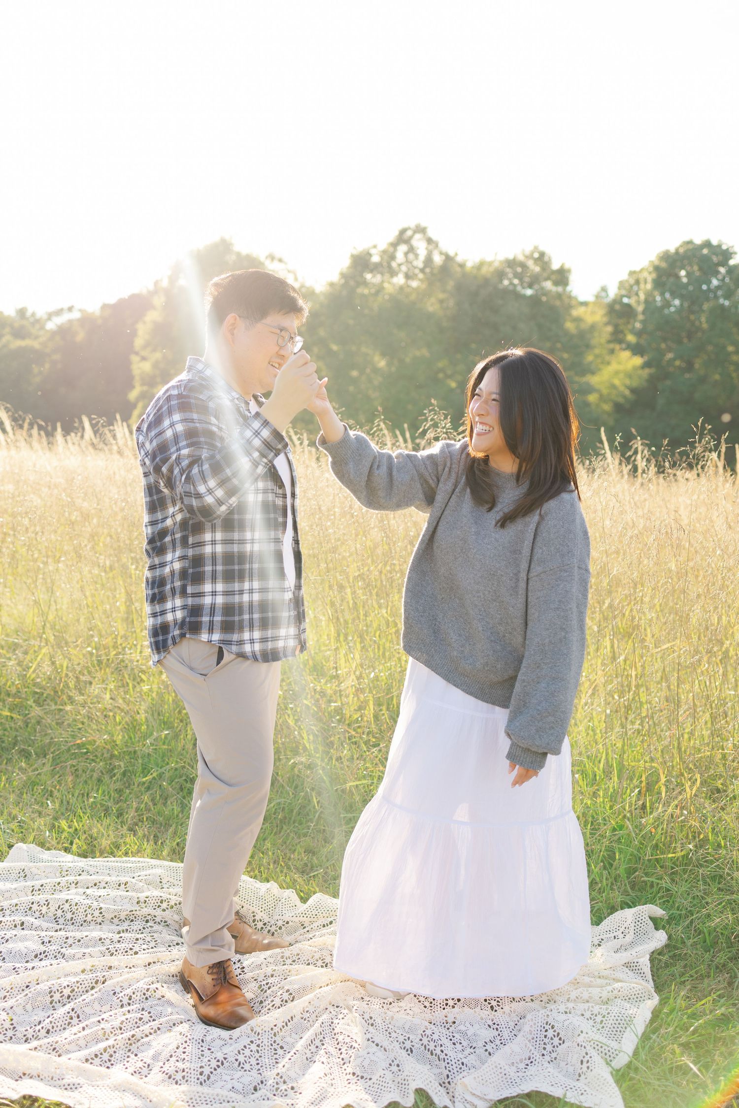 Couple sharing a joyful moment on a lace blanket in a sunlit meadow, woman in gray sweater and white skirt touching mans face.