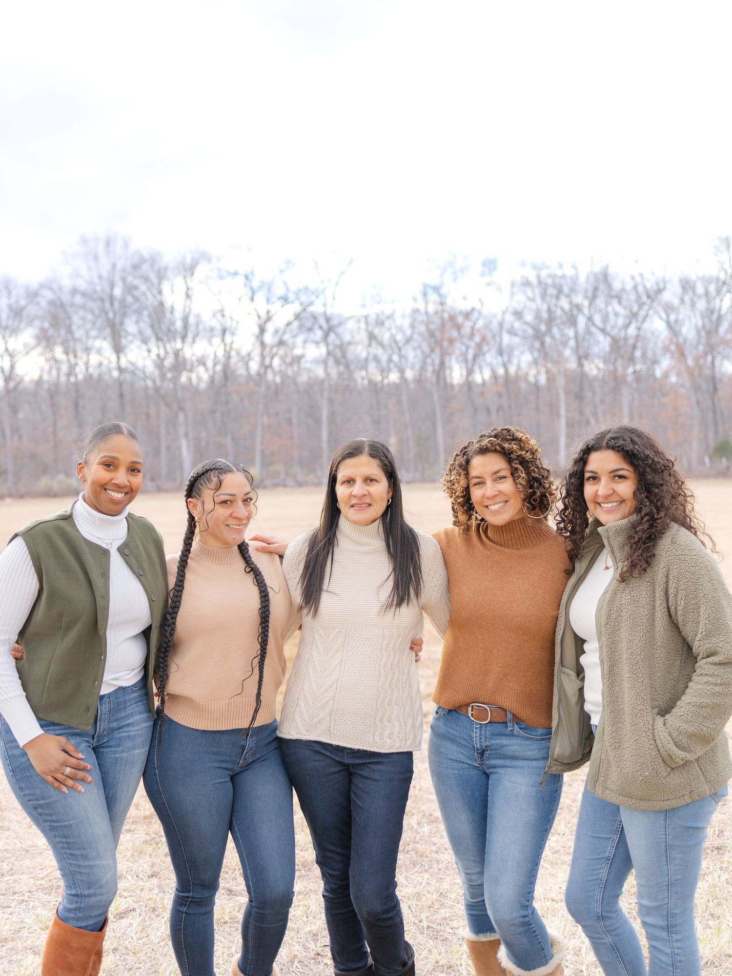 Five women smiling together outdoors in fall, wearing casual sweaters and jeans with bare trees in the background.