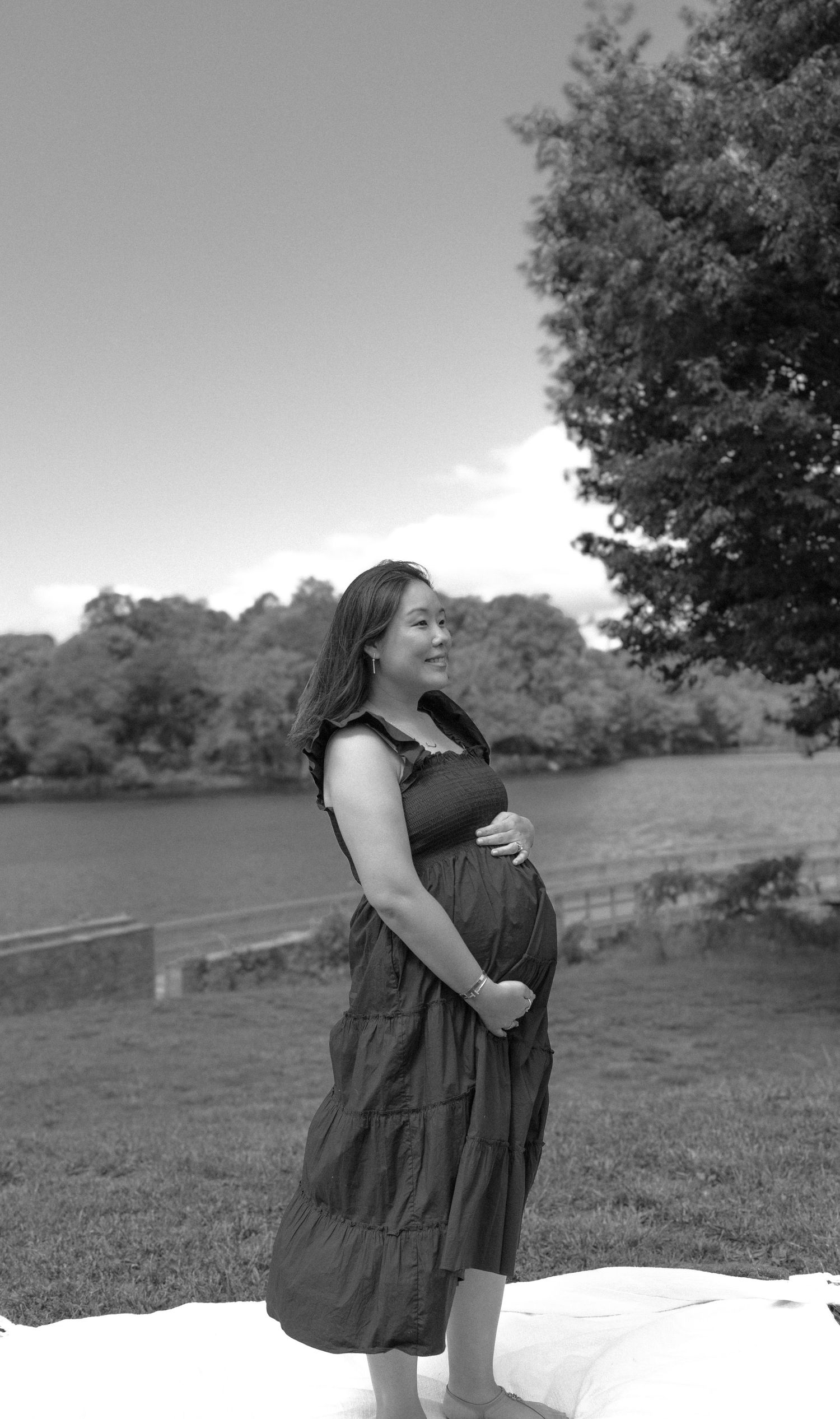 Black and white maternity photo of smiling pregnant woman in tiered dress outdoors by a lake surrounded by trees.
