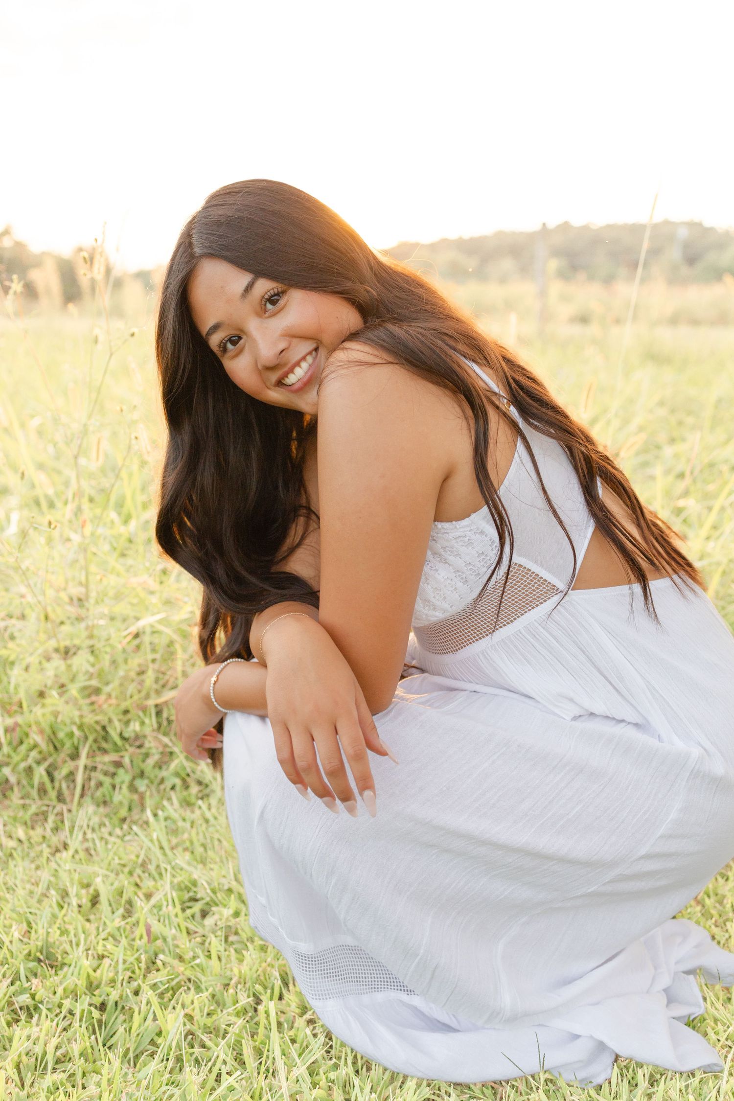Young woman in white dress crouching in a sunlit field, smiling warmly with long dark hair at golden hour.