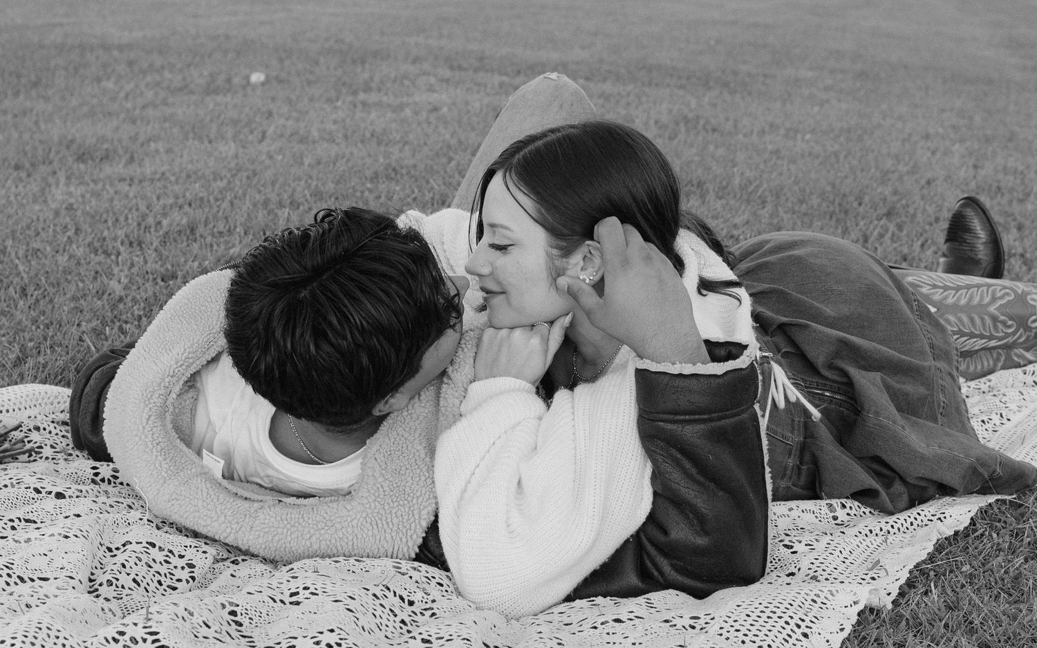 Black and white photo of a romantic couple lying on a blanket in a grassy field, smiling at each other.