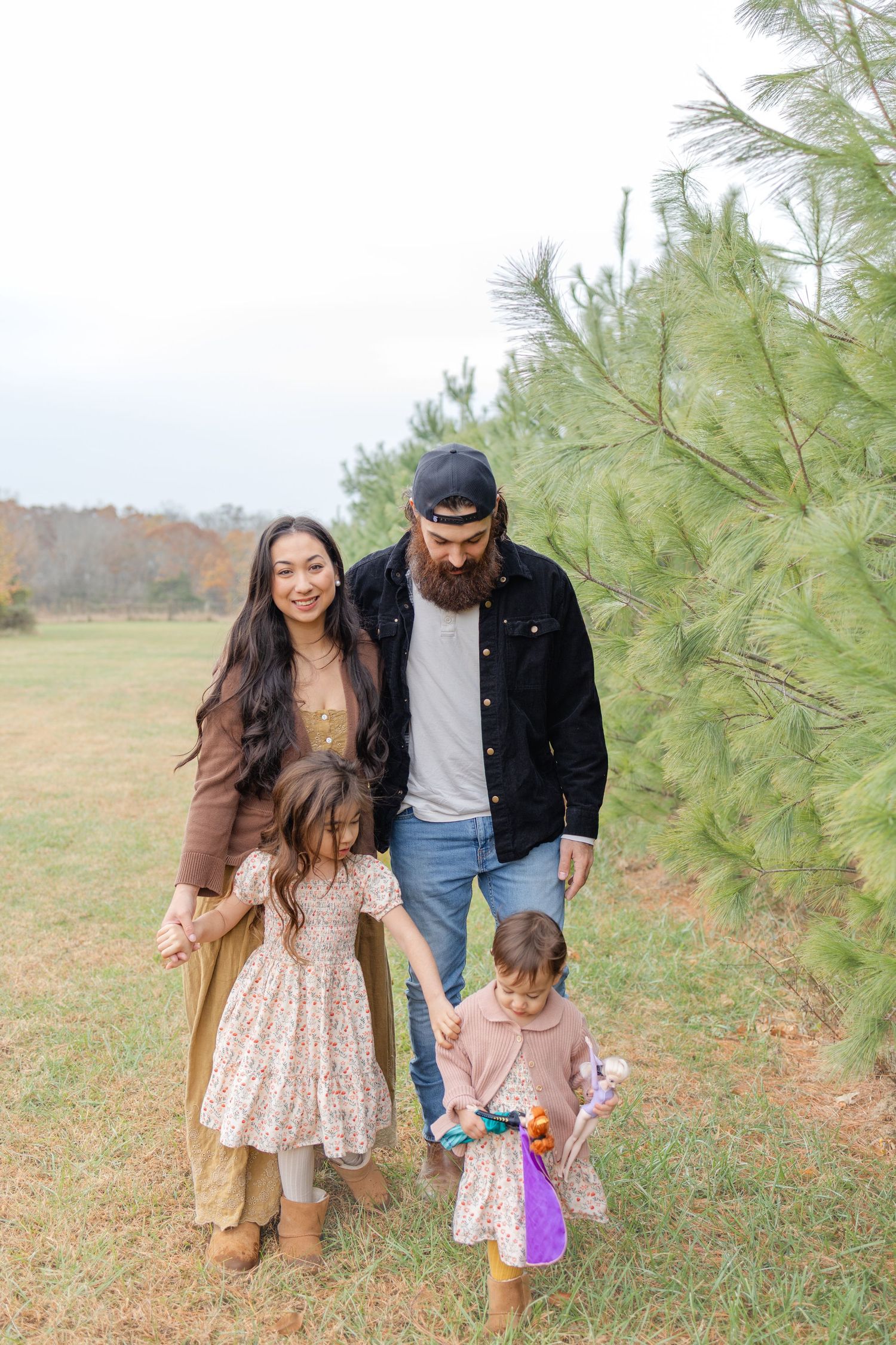 Family of four posing outdoors near pine trees in a field, parents holding hands with two young daughters.