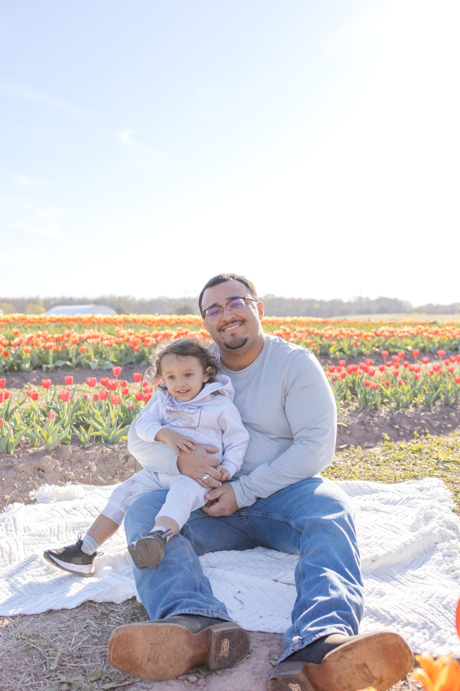A father and toddler sit on a blanket in a vibrant tulip field, smiling together on a sunny day.