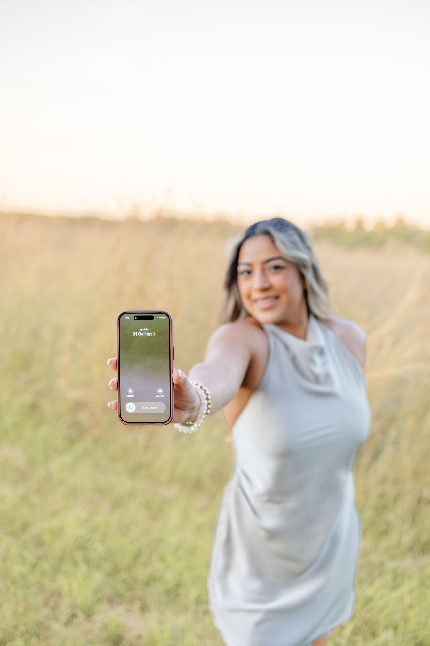 Young woman in a gray dress smiling in a golden field at sunset, holding an iPhone showing an incoming call screen.