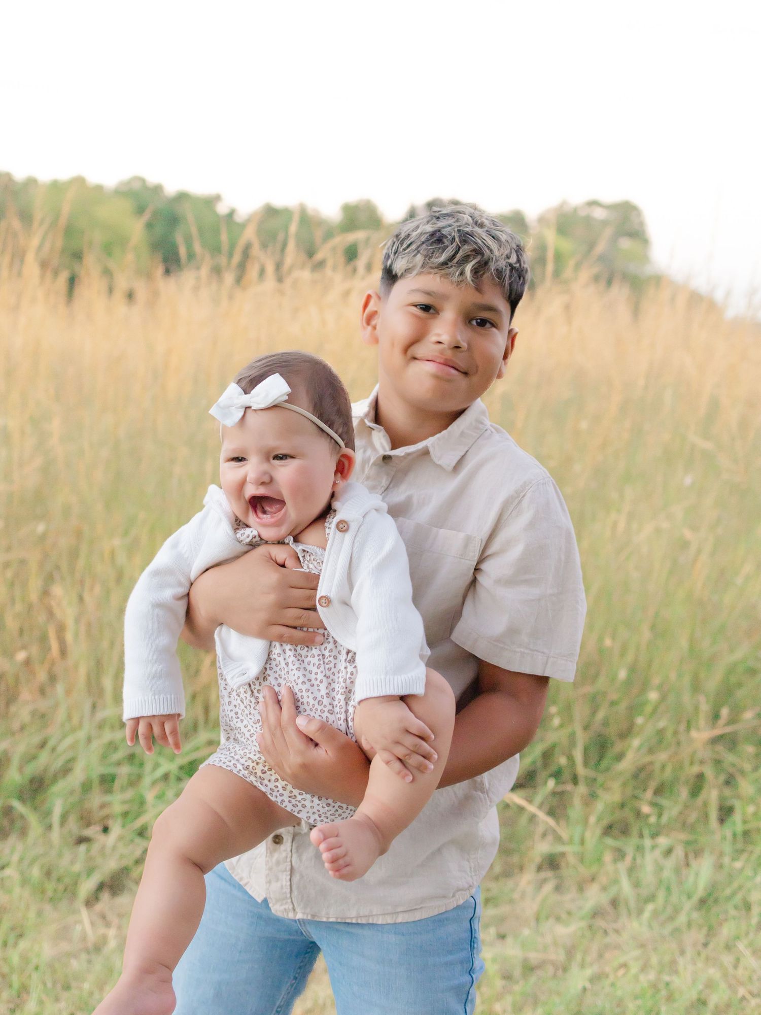 Older boy holding smiling baby girl in white bow in a golden field during an outdoor family photo session.