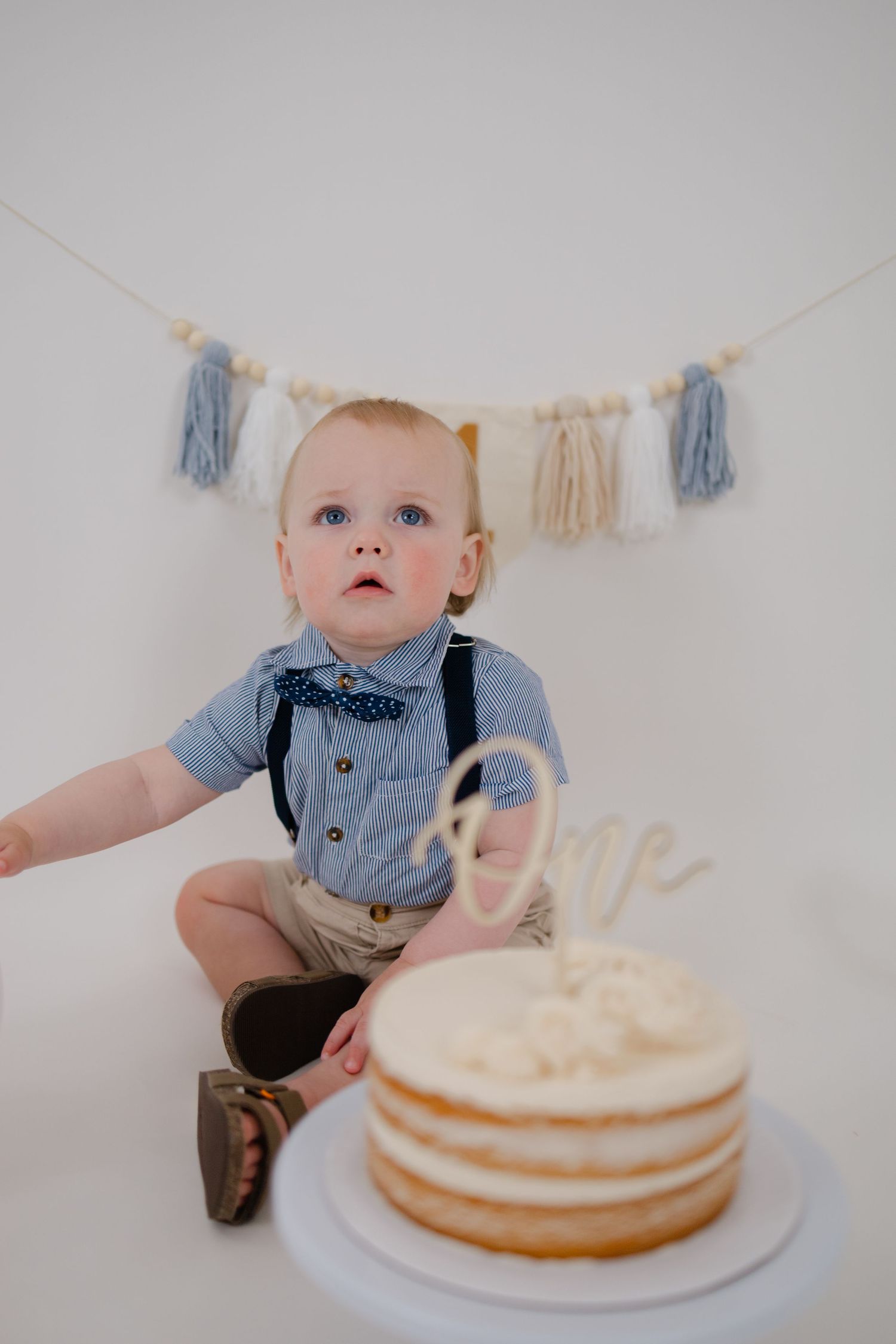 A baby in a blue shirt and suspenders sits near a white birthday cake with decorative tassels hanging on the wall.