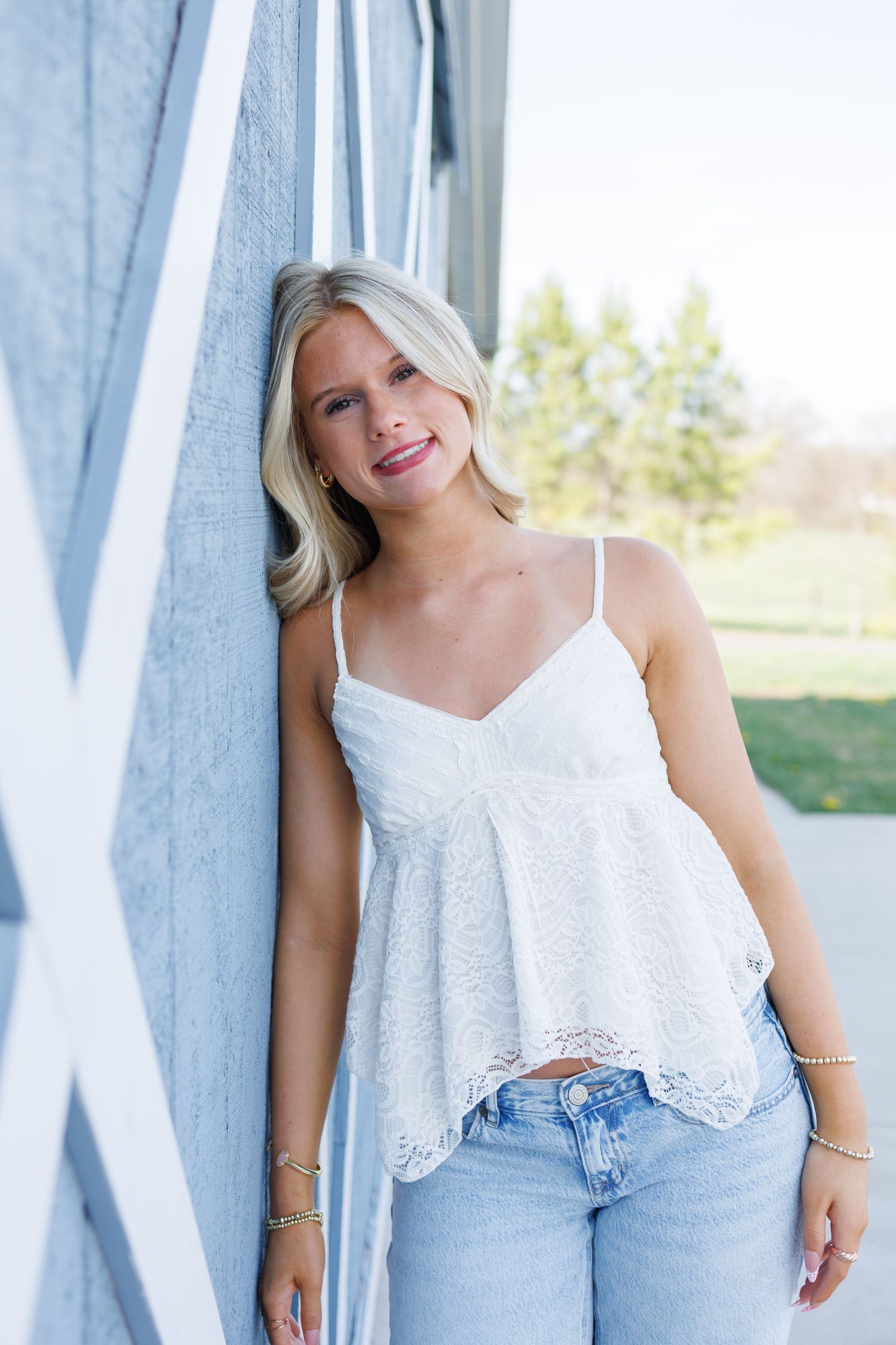 Young woman in white lace cami top and light wash jeans leaning against a blue barn wall outdoors.
