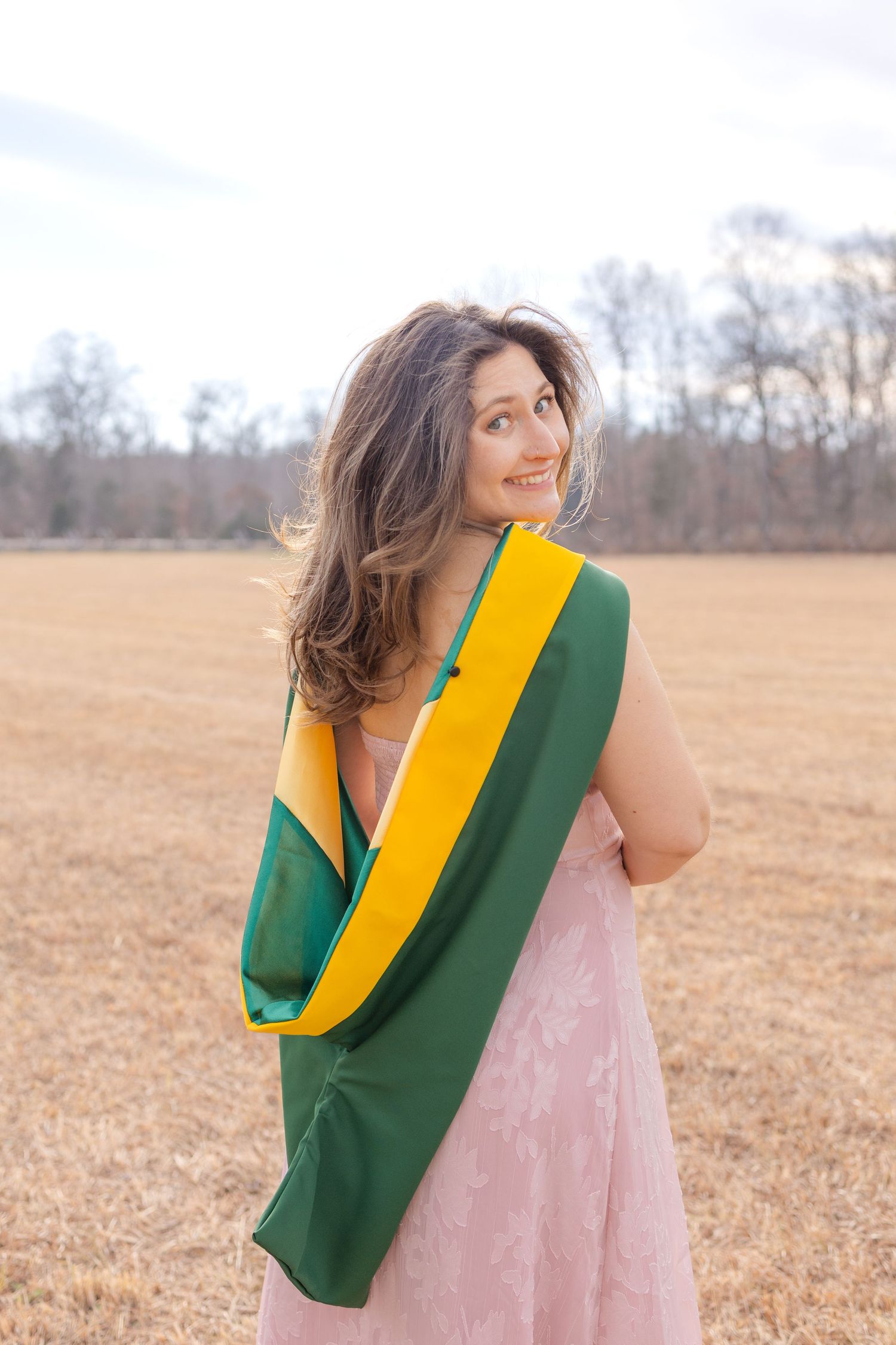 Smiling woman in pink dress wrapped in green and yellow flag standing in an open field with bare trees.