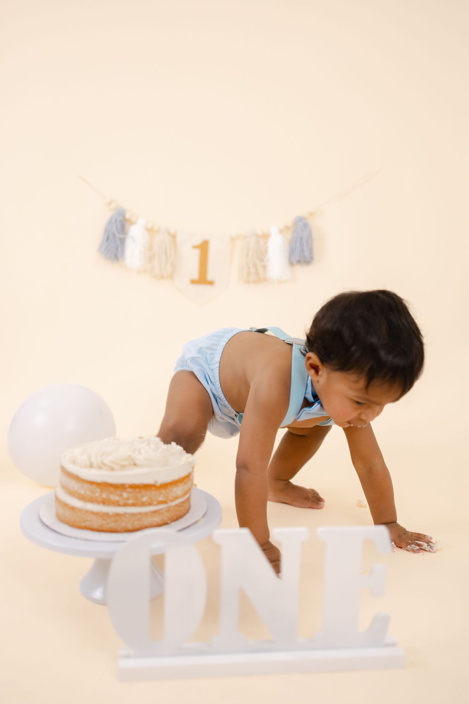 A baby in a white outfit crawls near a birthday cake during a first birthday cake smash photo session.
