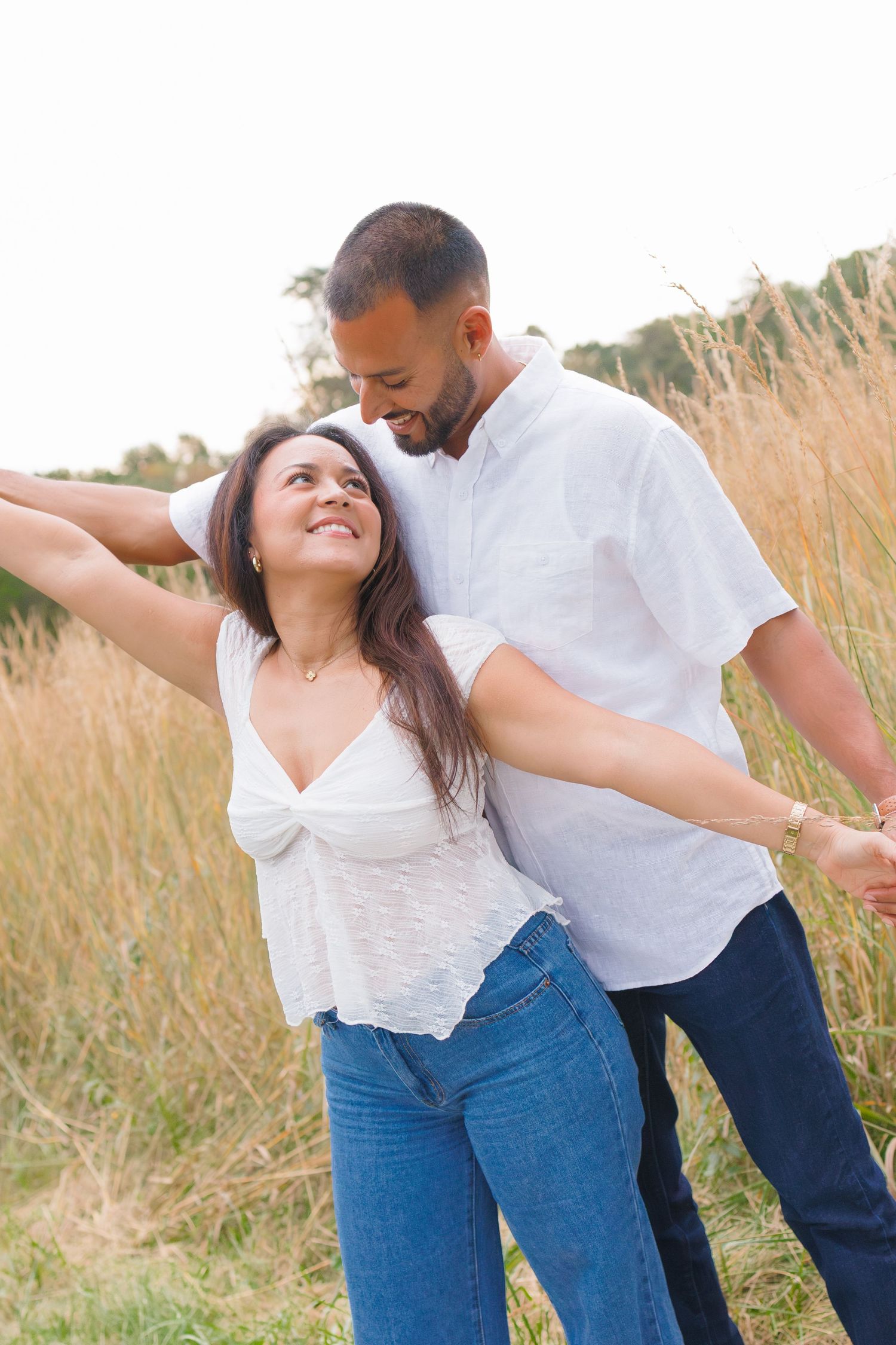 A couple in white tops and jeans dance joyfully in a golden field, sharing a tender smile.