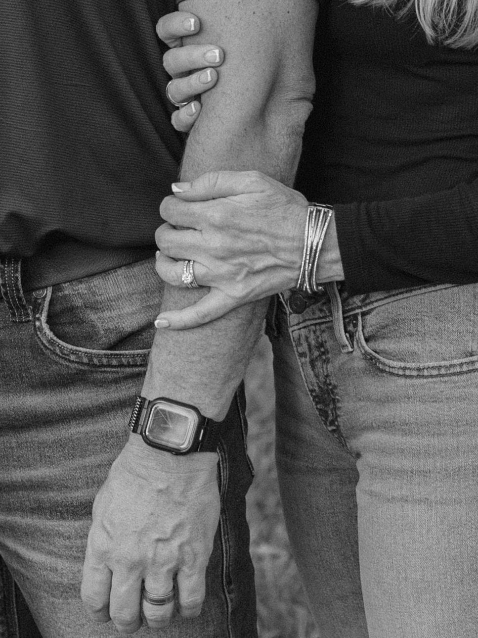Black and white photo of a couple holding hands, showing rings, a watch, and a bracelet, wearing jeans.