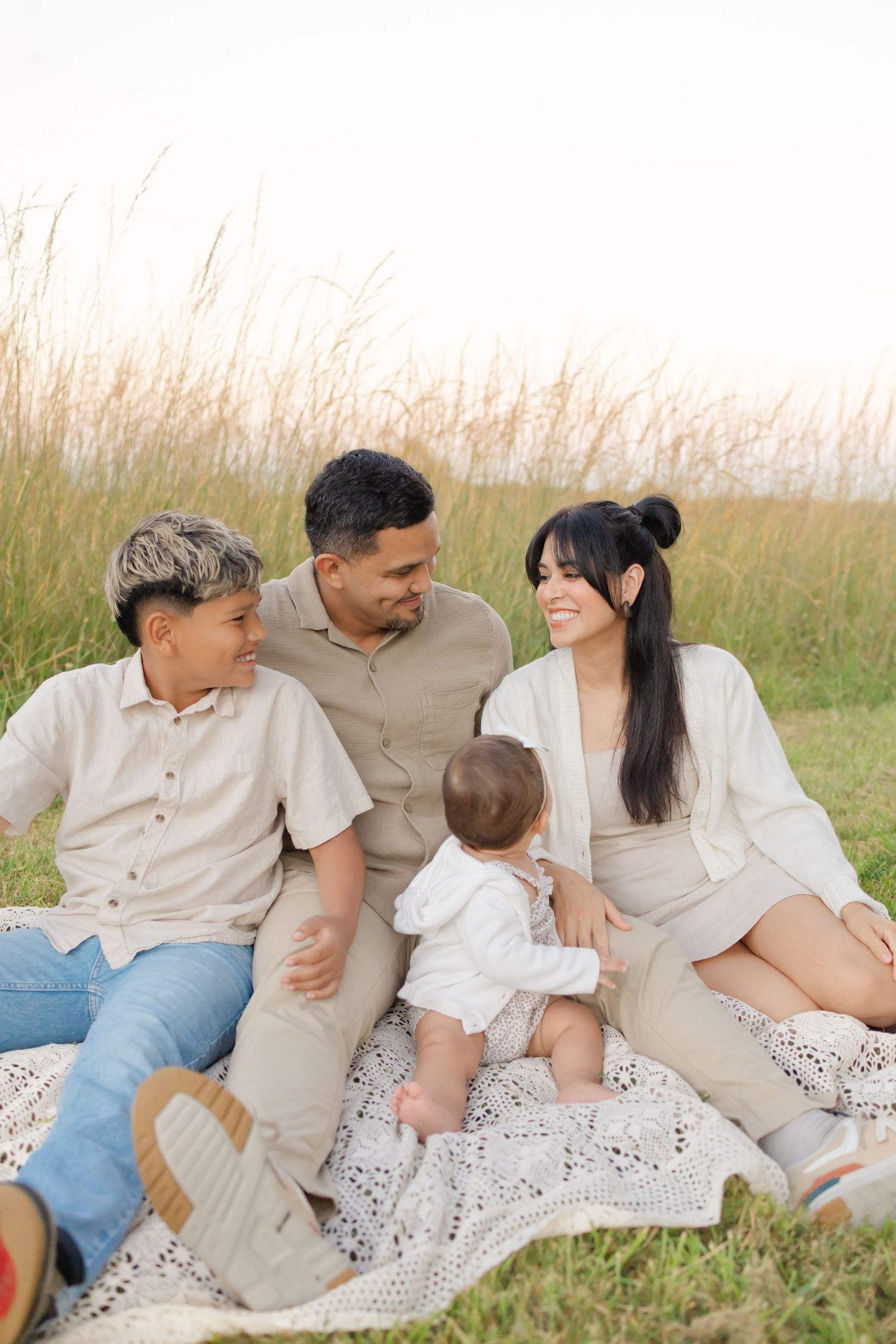 Family of four sitting on blanket in golden grass field, parents smiling at baby while older child looks on.
