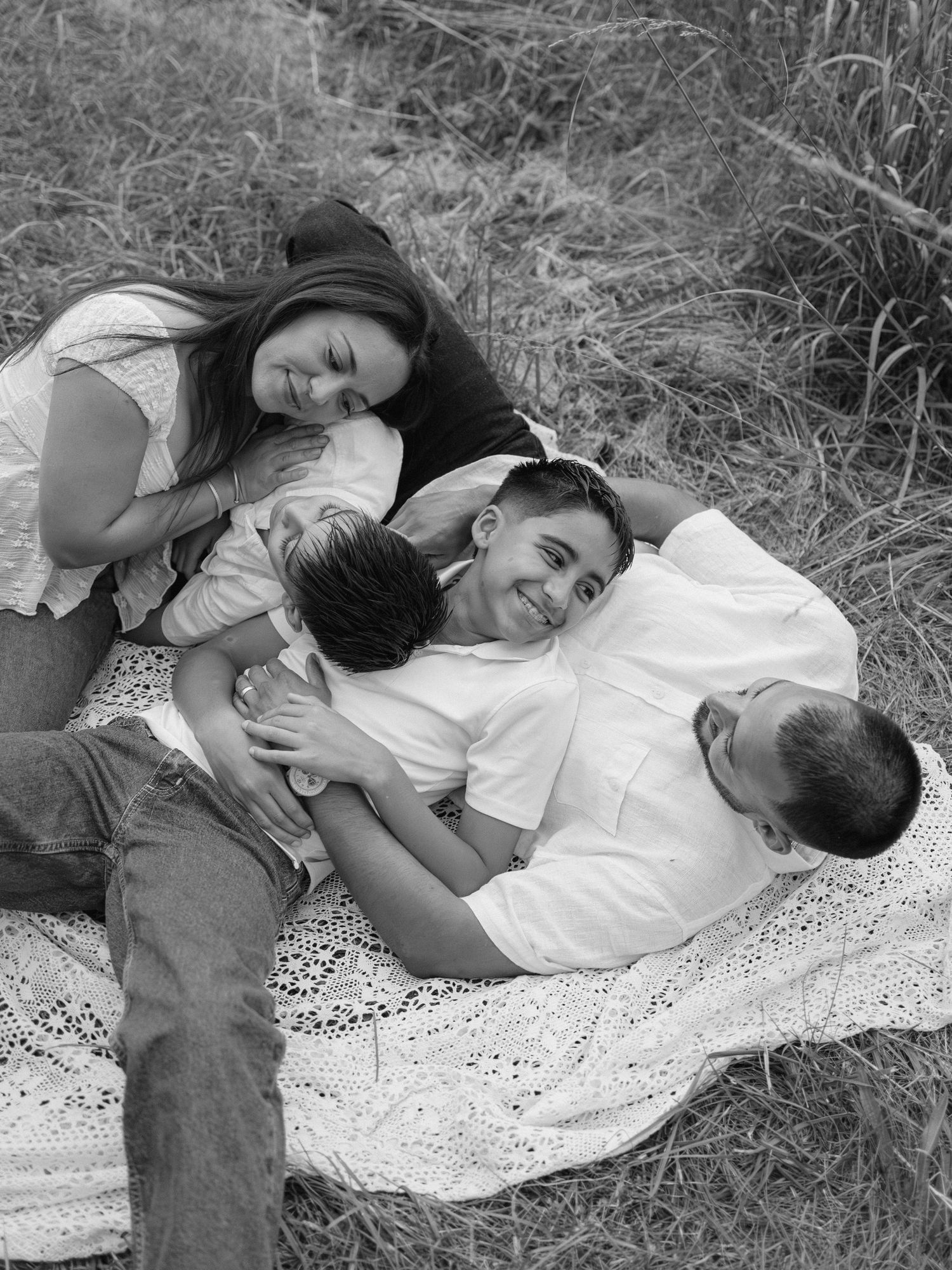 Black and white family photo of parents and two children laughing together on a blanket in a grassy field.