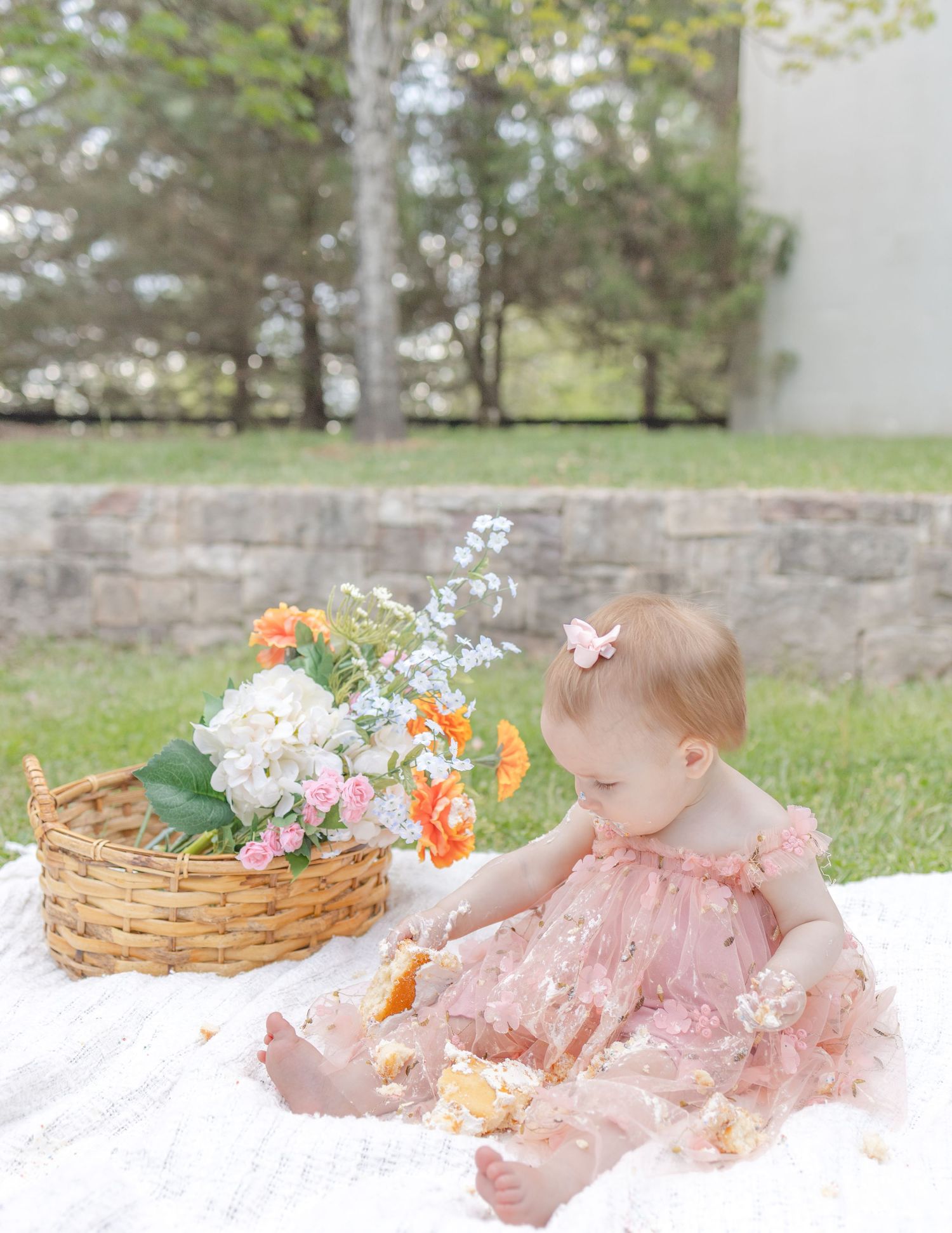 Baby girl in pink floral dress sits on white blanket outdoors, reaching toward a wicker basket filled with colorful spring flowers.